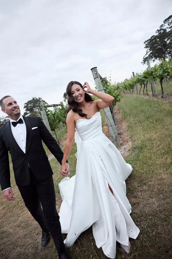 A smiling bride in a white strapless wedding gown holding hands with a groom in a black tuxedo with a bow tie, walking through a vineyard.