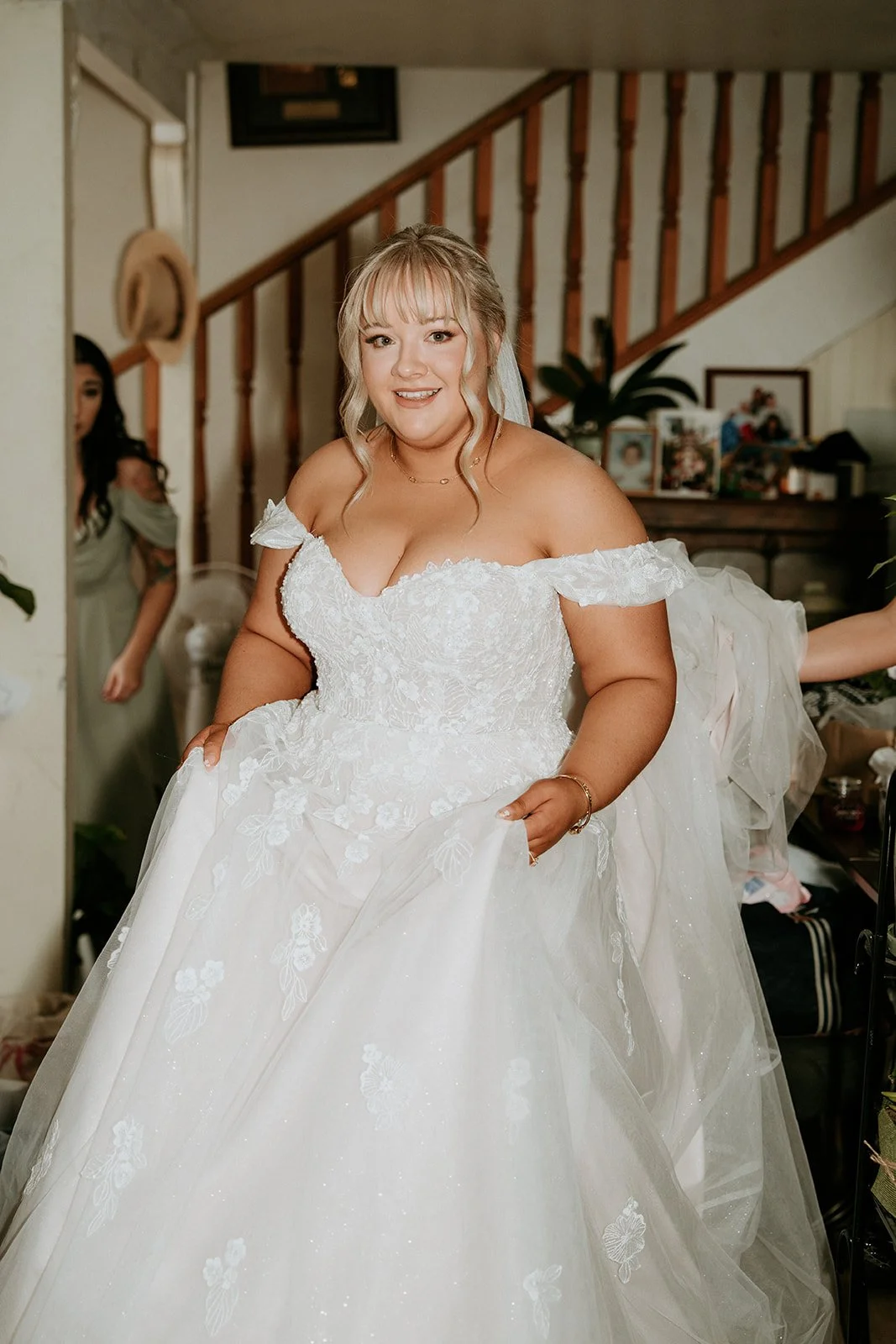 A smiling bride in a white off-the-shoulder wedding dress in a cozy indoor setting.