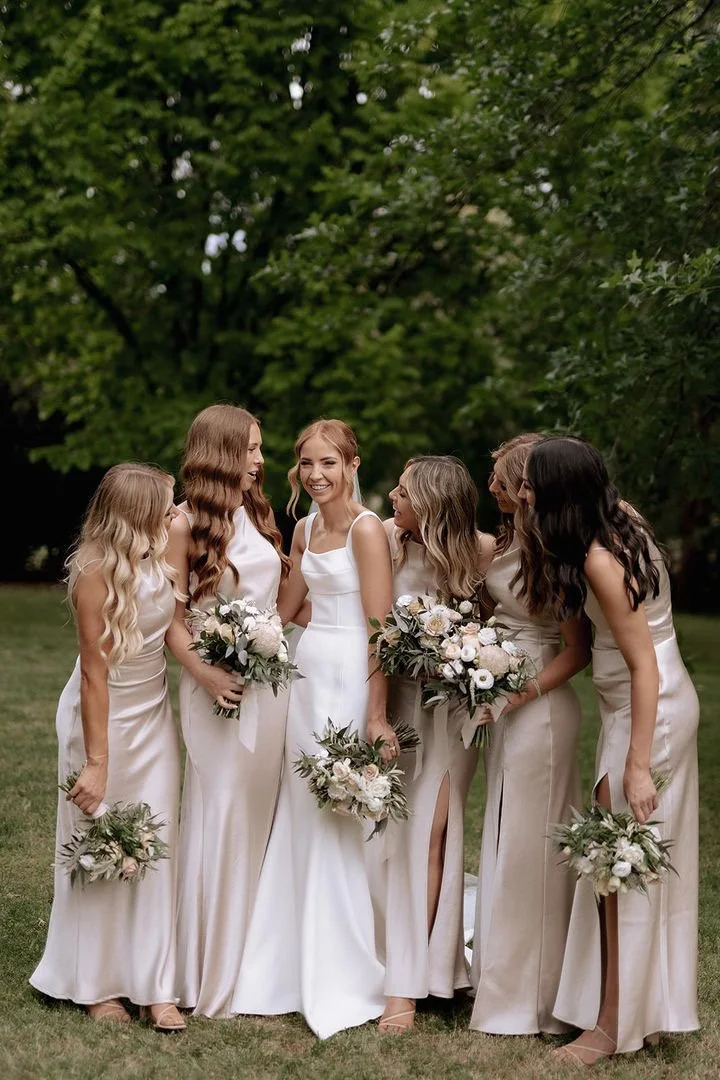 Group of six women at a wedding, wearing white dresses, holding bouquets of flowers, standing in a green outdoor setting, having a conversation, and smiling.