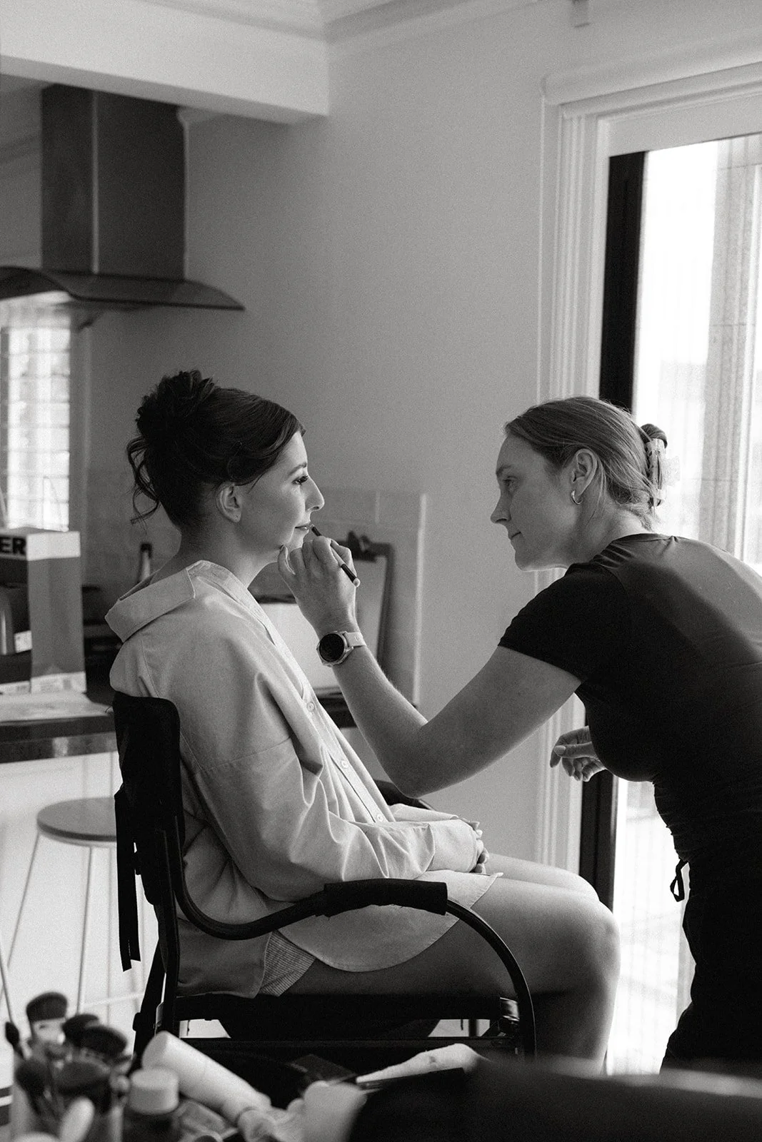 A woman applying makeup to another woman who is sitting in a chair, indoors near a window. The woman sitting is dressed casually, and the makeup artist is leaning in, smiling.