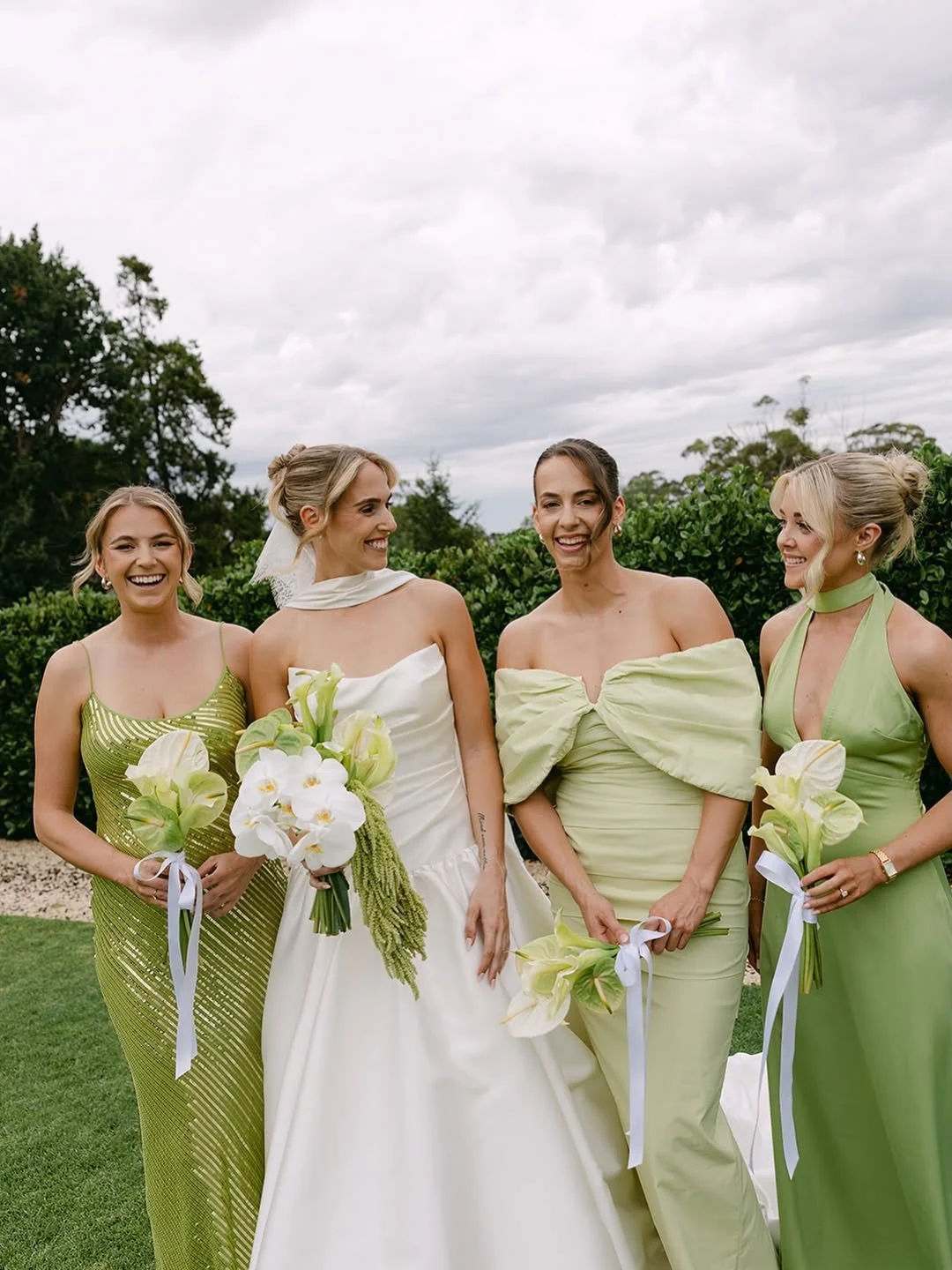 Four women in elegant dresses standing outdoors, smiling and holding bouquets of white flowers, with a cloudy sky and greenery in the background.