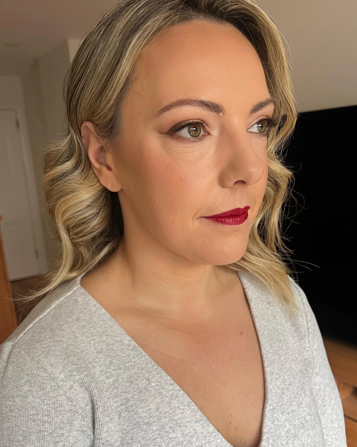 A woman with blonde, wavy hair, light skin, green eyes, wearing red lipstick and a gray shirt, looking slightly to the right.