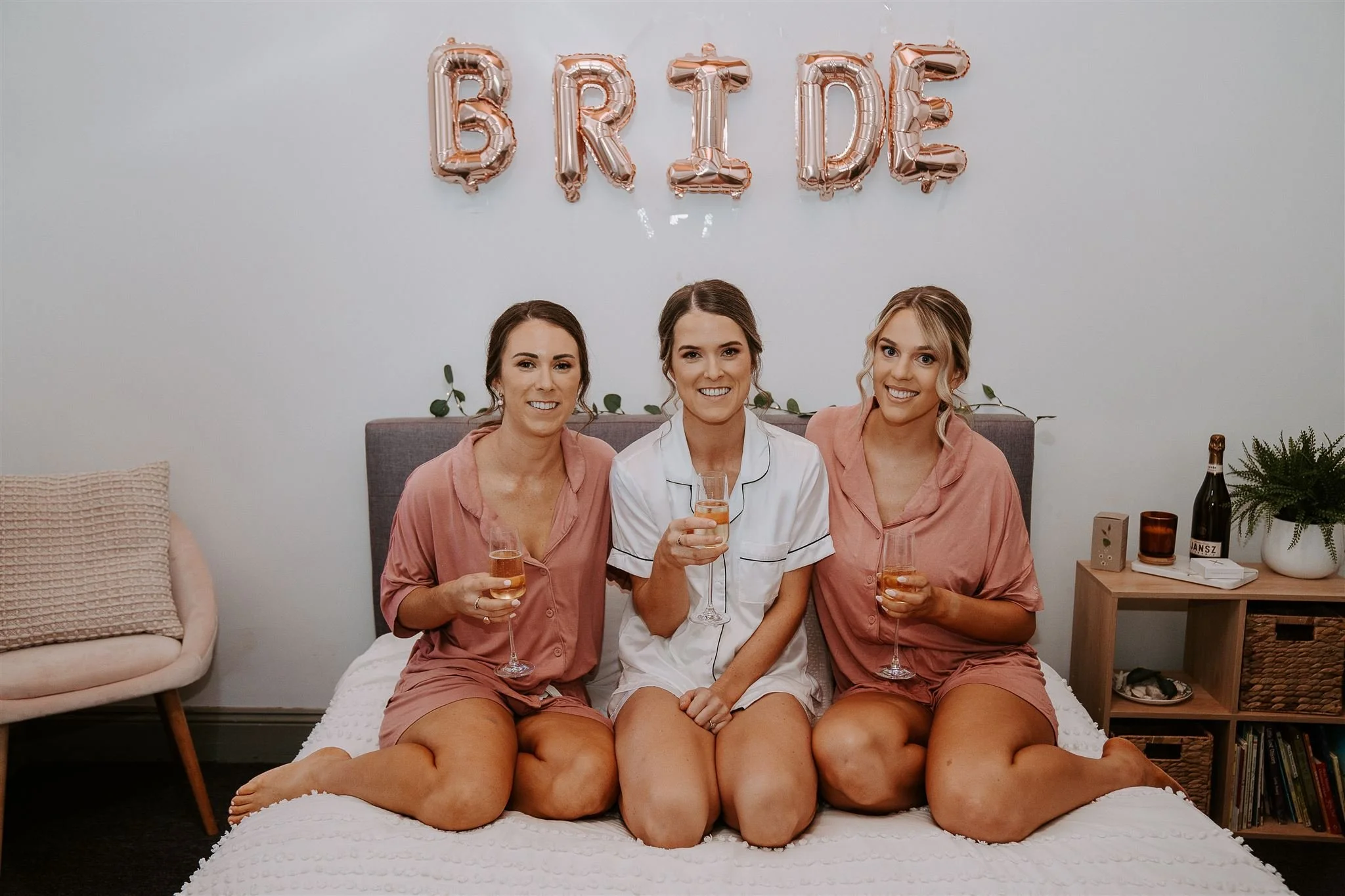 Three women in pajamas sitting on a bed with champagne glasses, celebrating a bridal event with a "BRIDE" balloon banner in the background.