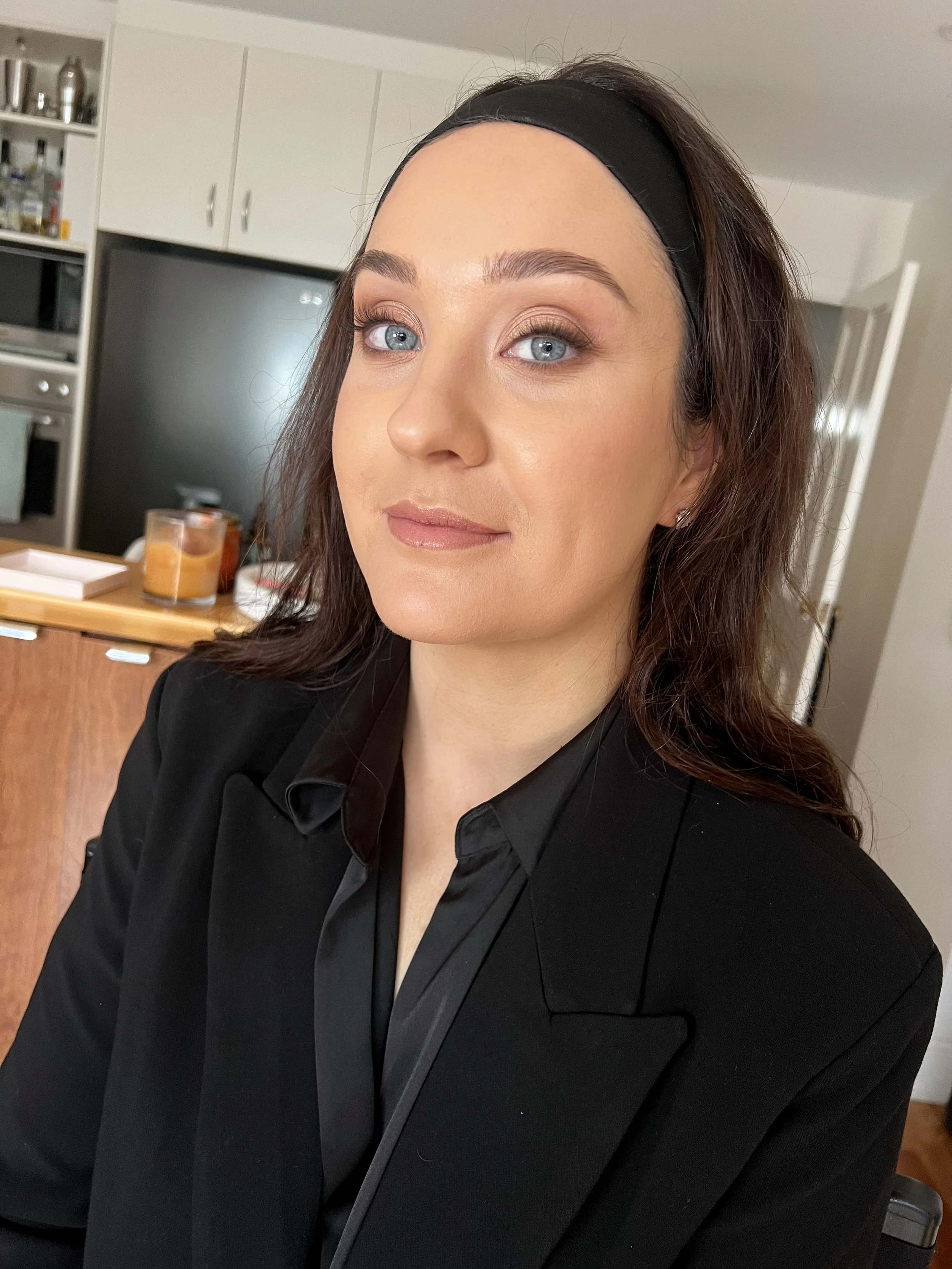 A woman with blue eyes and brown hair wearing a black blazer and headband, posing in a kitchen.