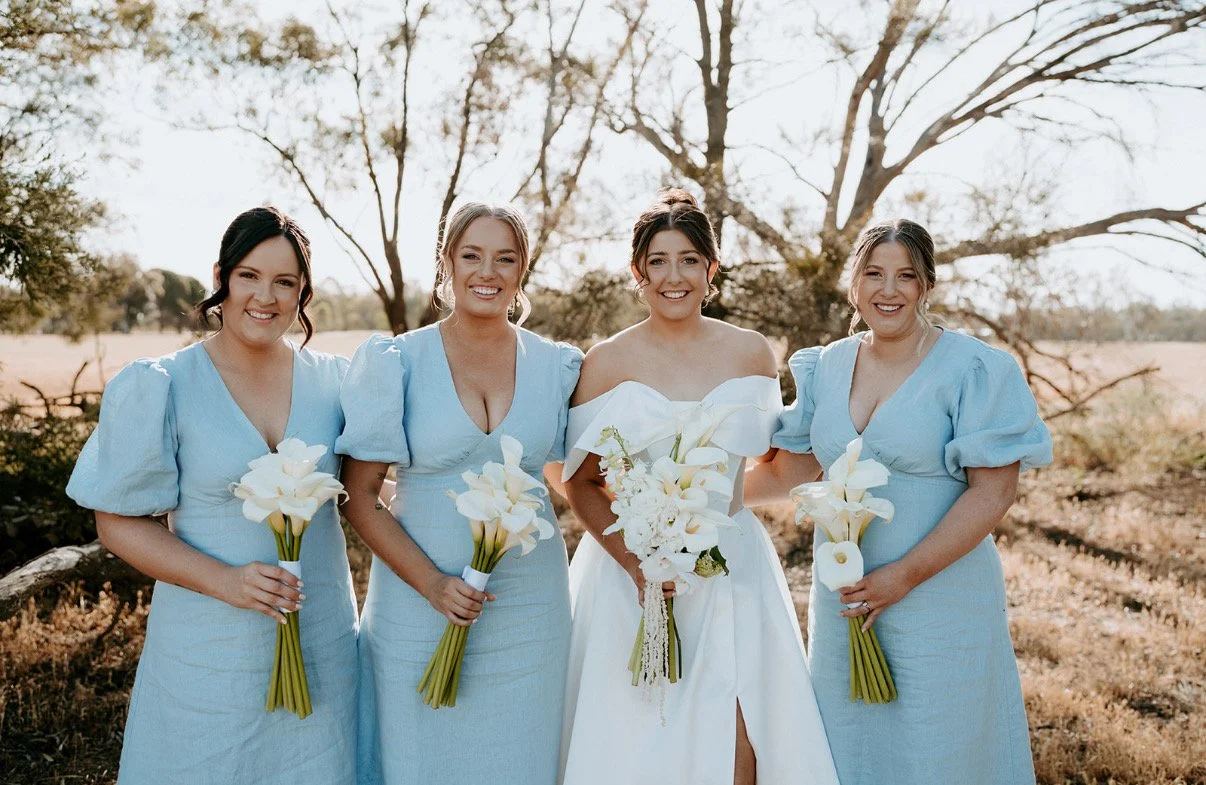 Four women, one dressed as a bride in a white wedding gown and three bridesmaids in matching light blue dresses, standing outdoors with trees in the background. They are holding bouquets of white flowers and smiling at the camera.
