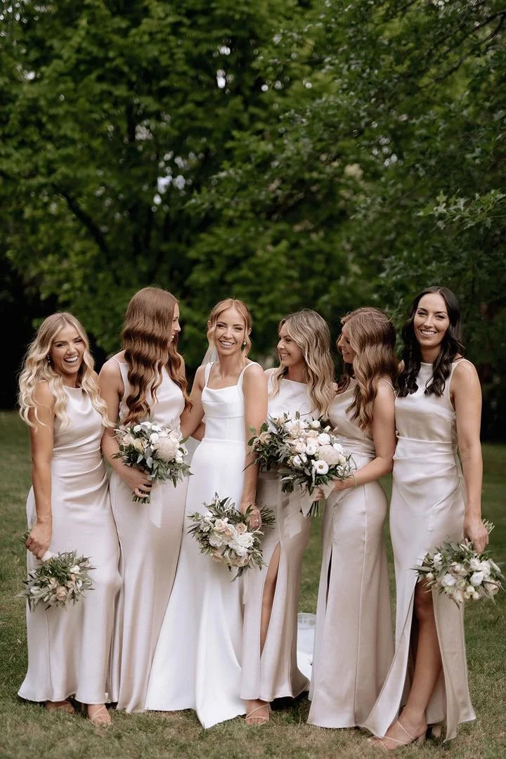 Six women in white dresses holding bouquets, standing outdoors on grass with green trees in the background, smiling and celebrating.
