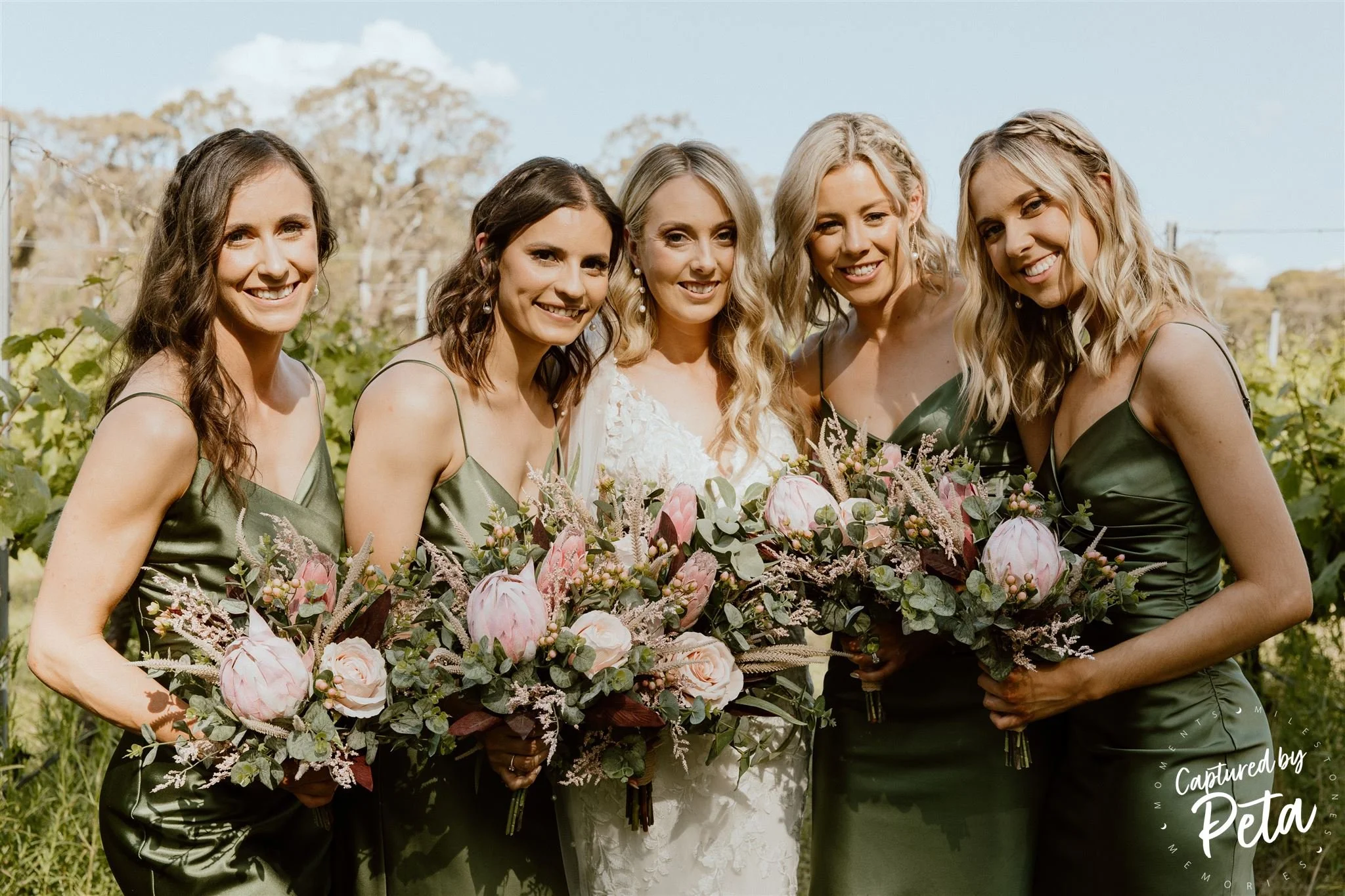 Group of five women at a wedding, holding bouquets of pink and white flowers, outdoors on a sunny day with blue sky and trees in the background.