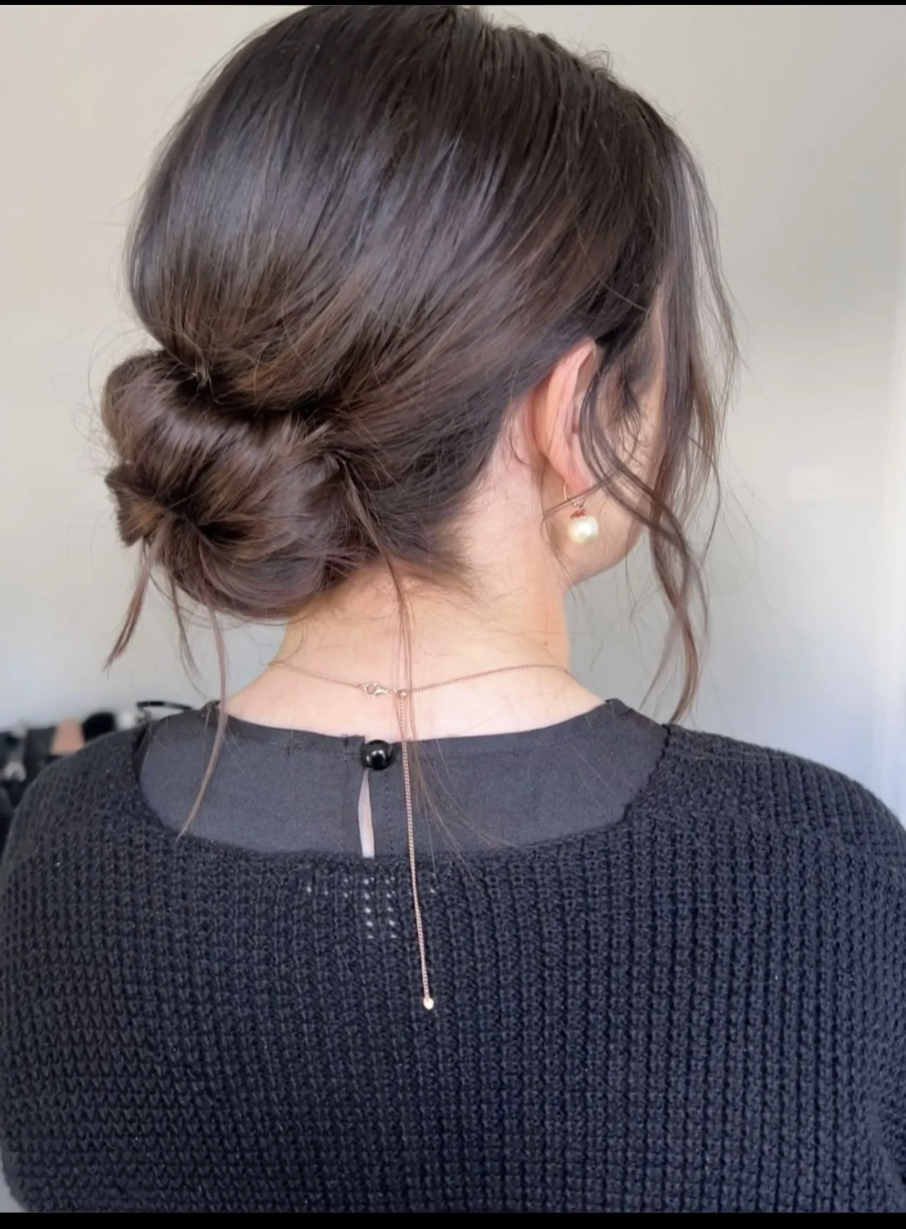Back view of a woman with dark brown hair styled in a loose bun, wearing pearl earrings and layered necklaces, and dressed in a black textured top.