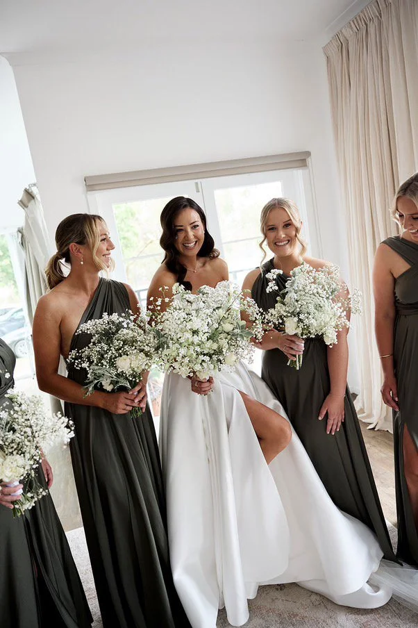 Bride in white wedding gown with four bridesmaids in dark gray dresses holding white flower bouquets, smiling inside a room with large windows and cream curtains.