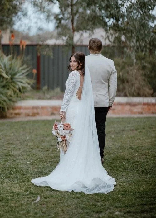 Bride in a white wedding dress holding a bouquet of pink and white flowers, standing on grass with a groom in a light gray suit, seen from the back outdoors.