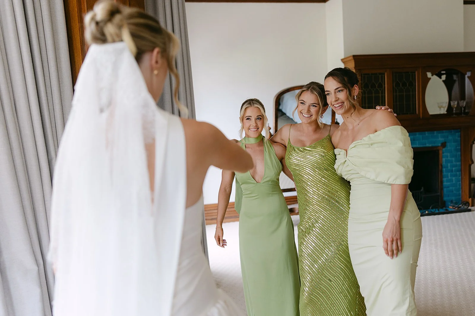 A bride with a wedding veil is looking at three women in elegant dresses in a hotel room, smiling and posing for a photo.