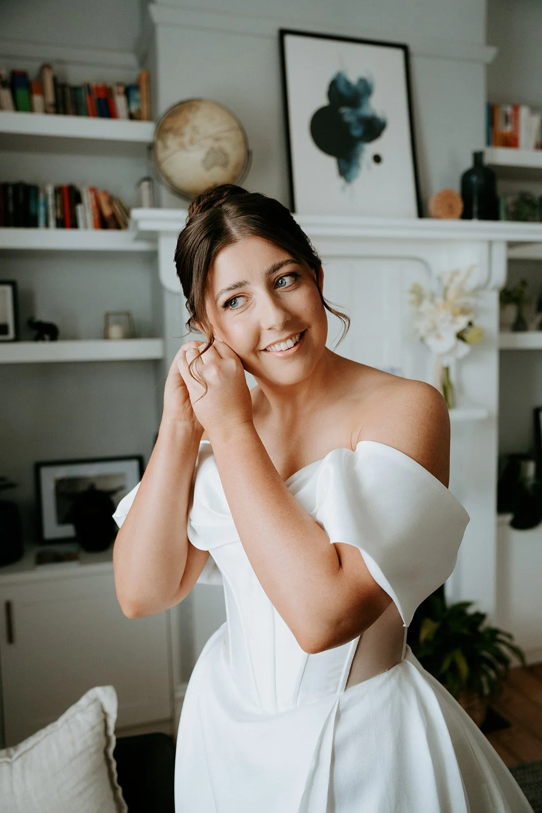 A woman in a white wedding dress smiling and adjusting her earring in a cozy, decorated room.