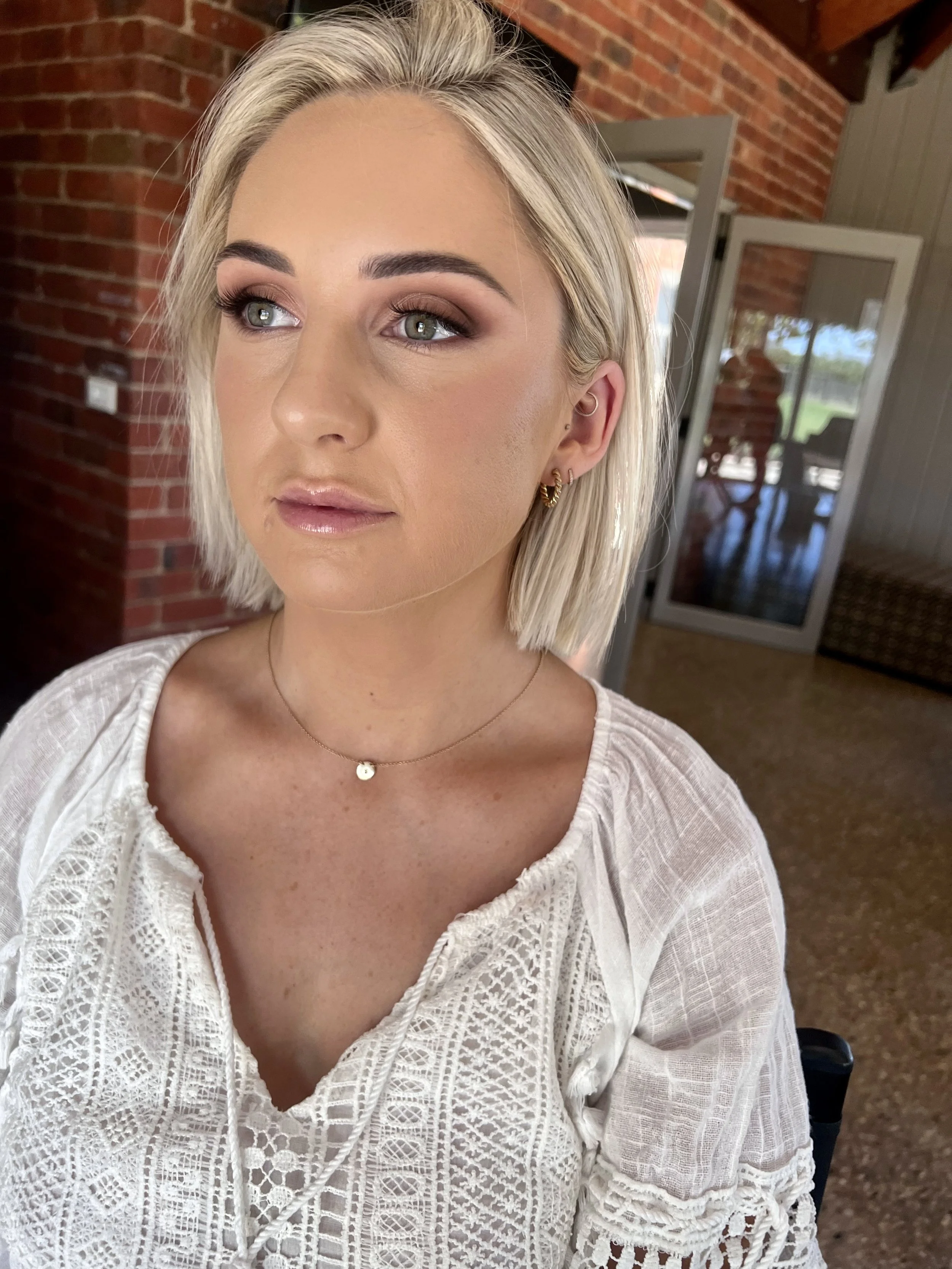 Close-up of a woman with short blonde hair and makeup, wearing a white lace top, a delicate necklace, and small gold hoop earrings, indoors with brick and wooden walls, a door, and a distant reflection in glass behind her.