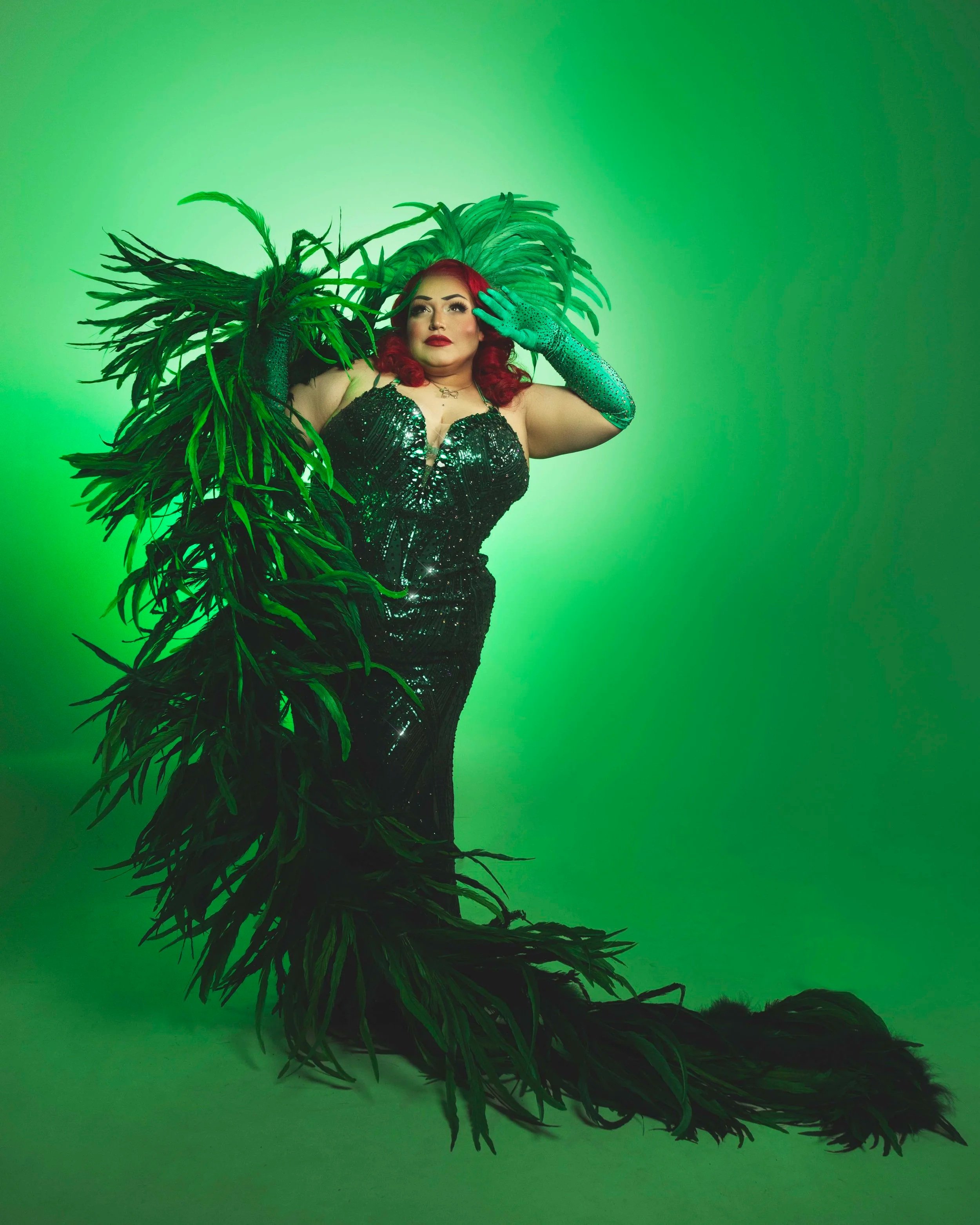 Bella Sin wears an emerald green, rhinestoned gown, feather boa and headpiece, and stands in front of a bright green backdrop
