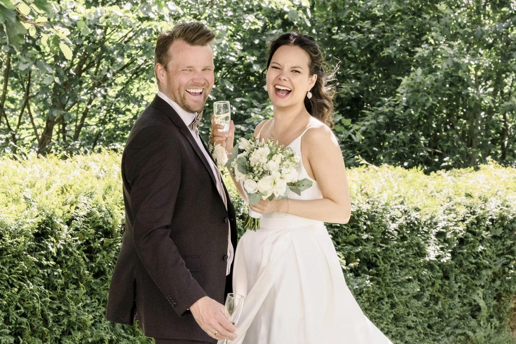 Et lykkelig brudepar smiler og ler utendørs, med mannen i svart dress og kvinnen i hvit brudekjole, begge holder toastglass og kvinnen holder en brudebukett