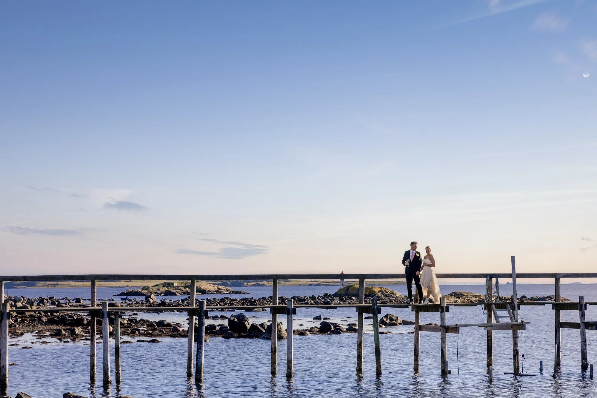 En brud og en brudgom står på en brygge langs sjøen under en klar himmel med fullmåne i bakgrunnen.