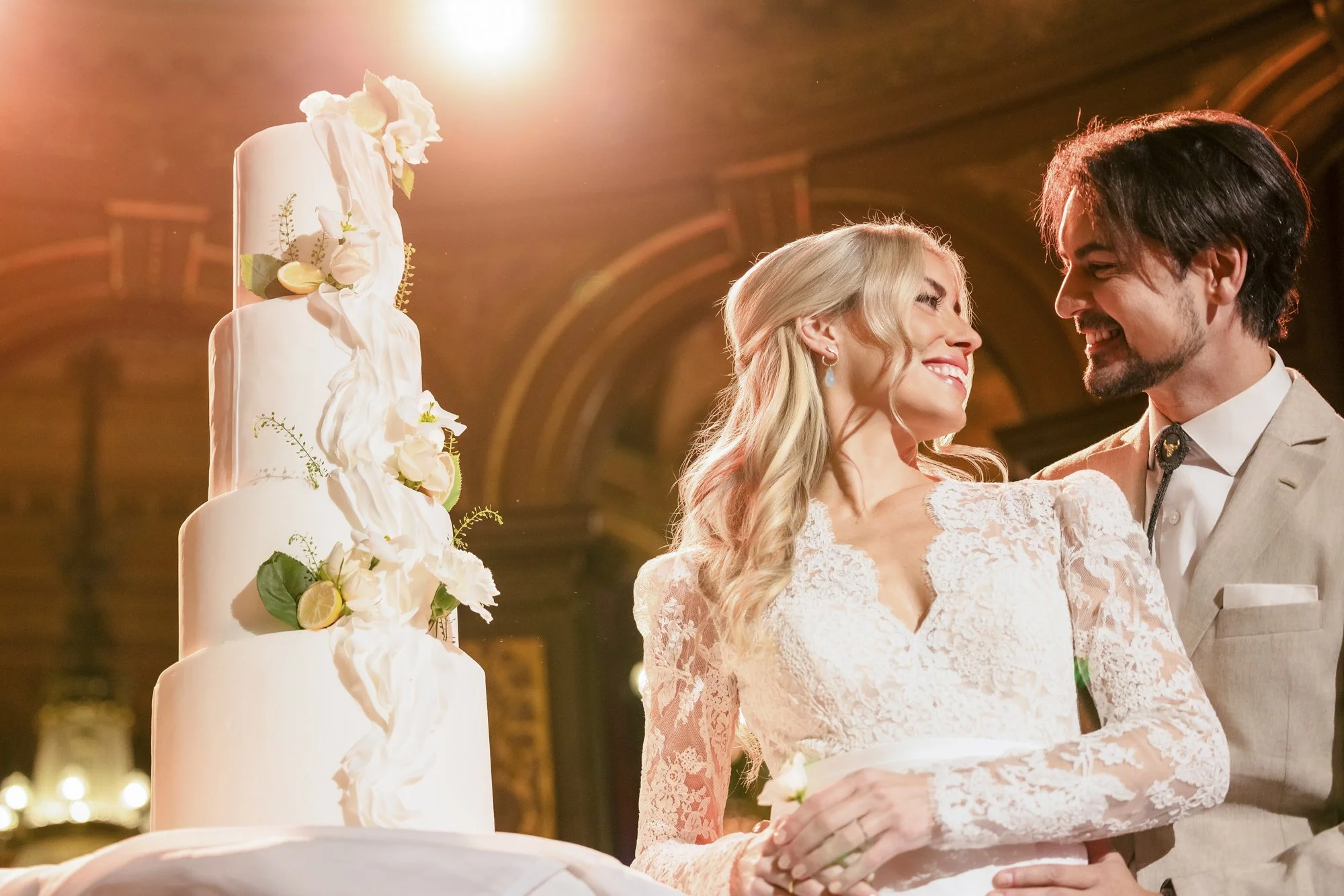 En nygift brud og brudgom smiler mot hverandre foran en bryllag cake i et vakkert, opplyst rom.