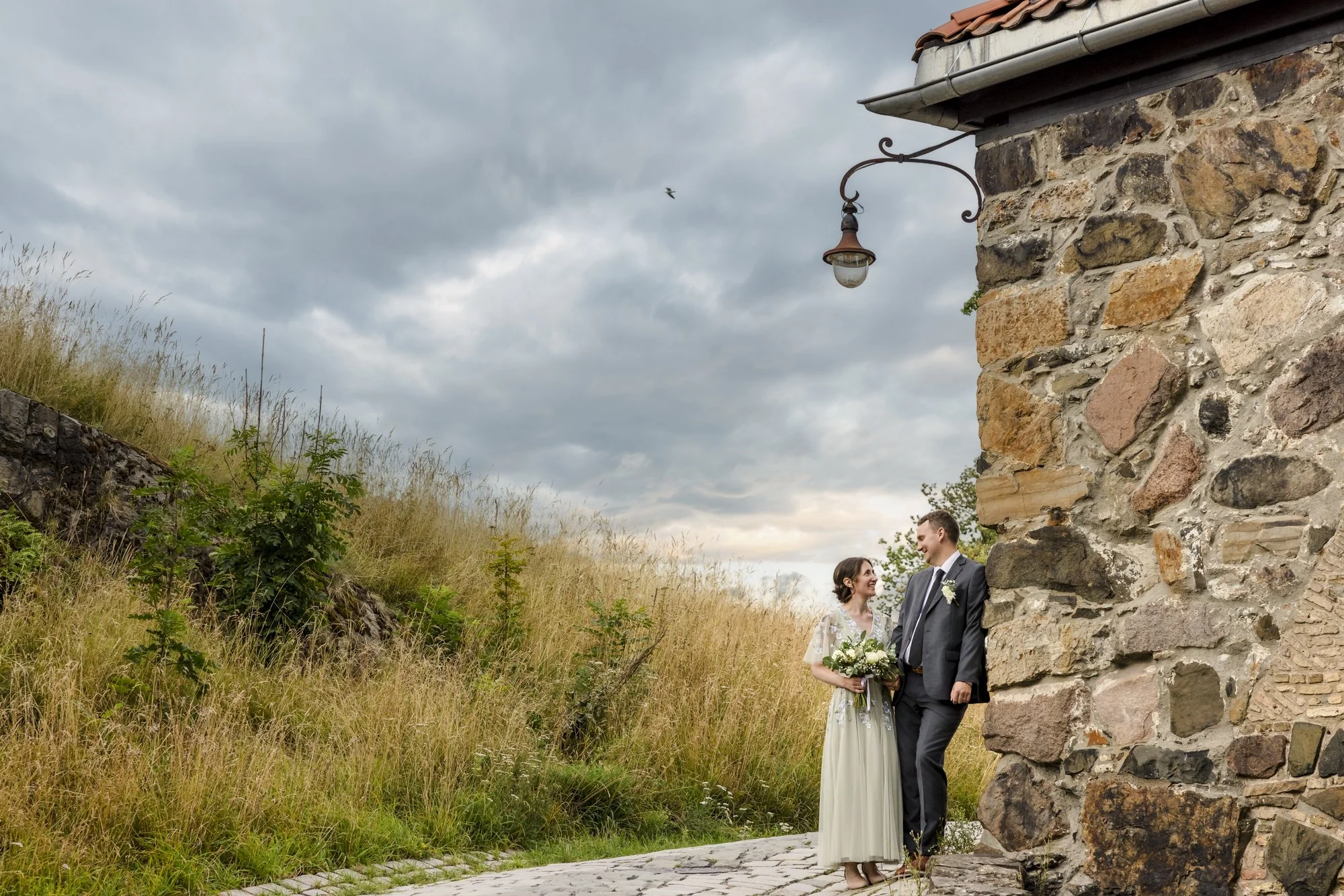 Et nygift par står sammen utendørs ved en gammel steinmur, med den vakre naturen og en mørk himmel i bakgrunnen.