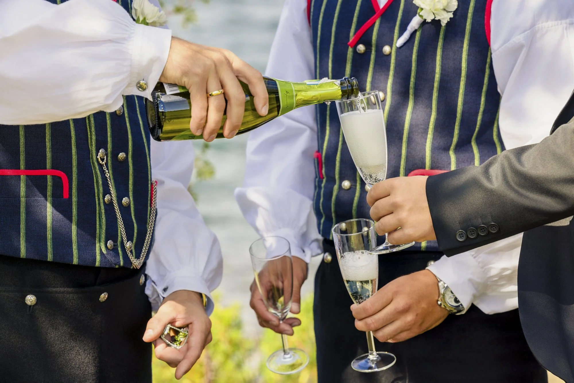 Personer kledd i tradisjonelle klær serverer og mottar champagne i glass ved utendørs sammenkomst.