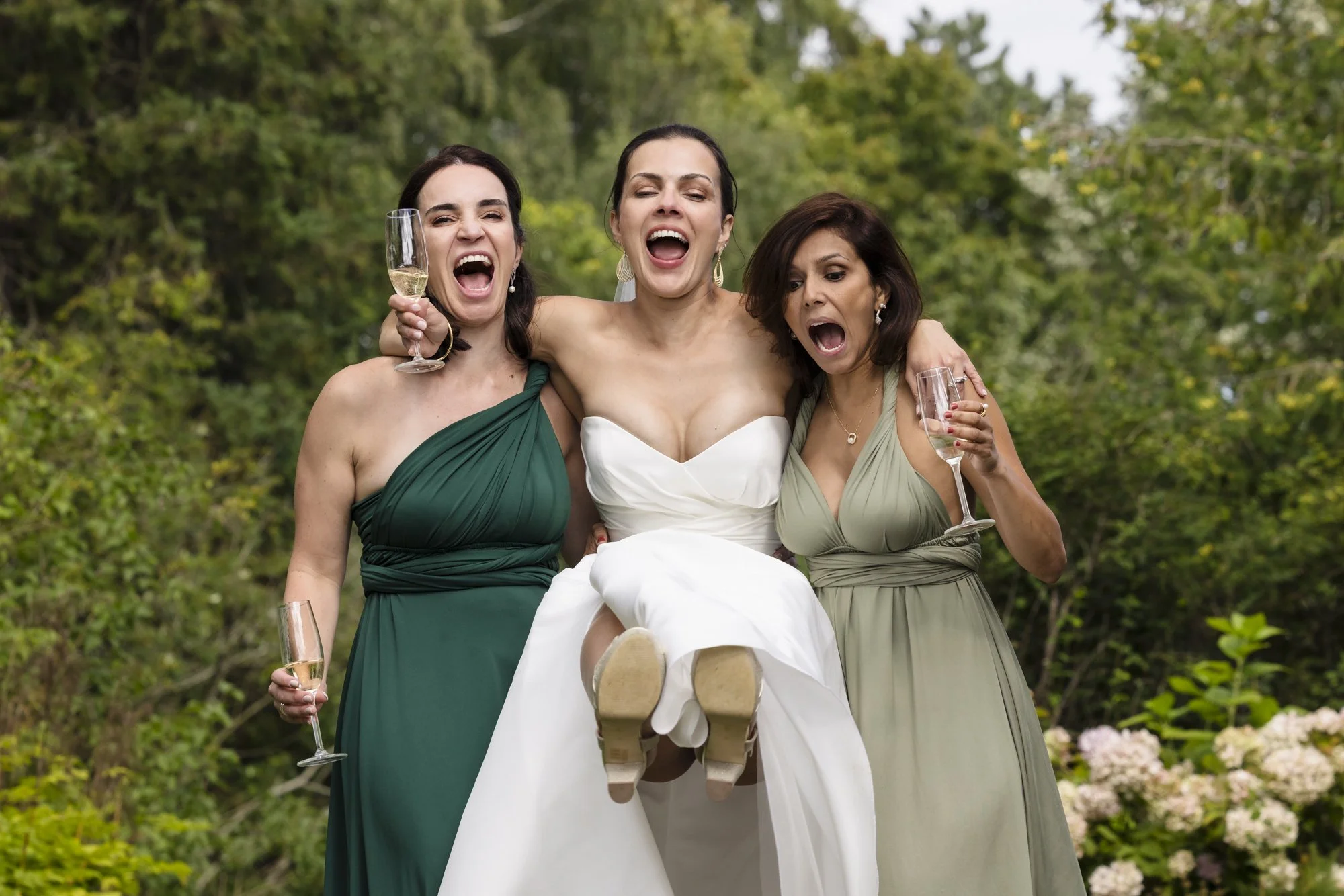 Tre kvinner i kjoler feirer med champagne utendørs, en av dem er i brudekjole, og de ser glade ut.