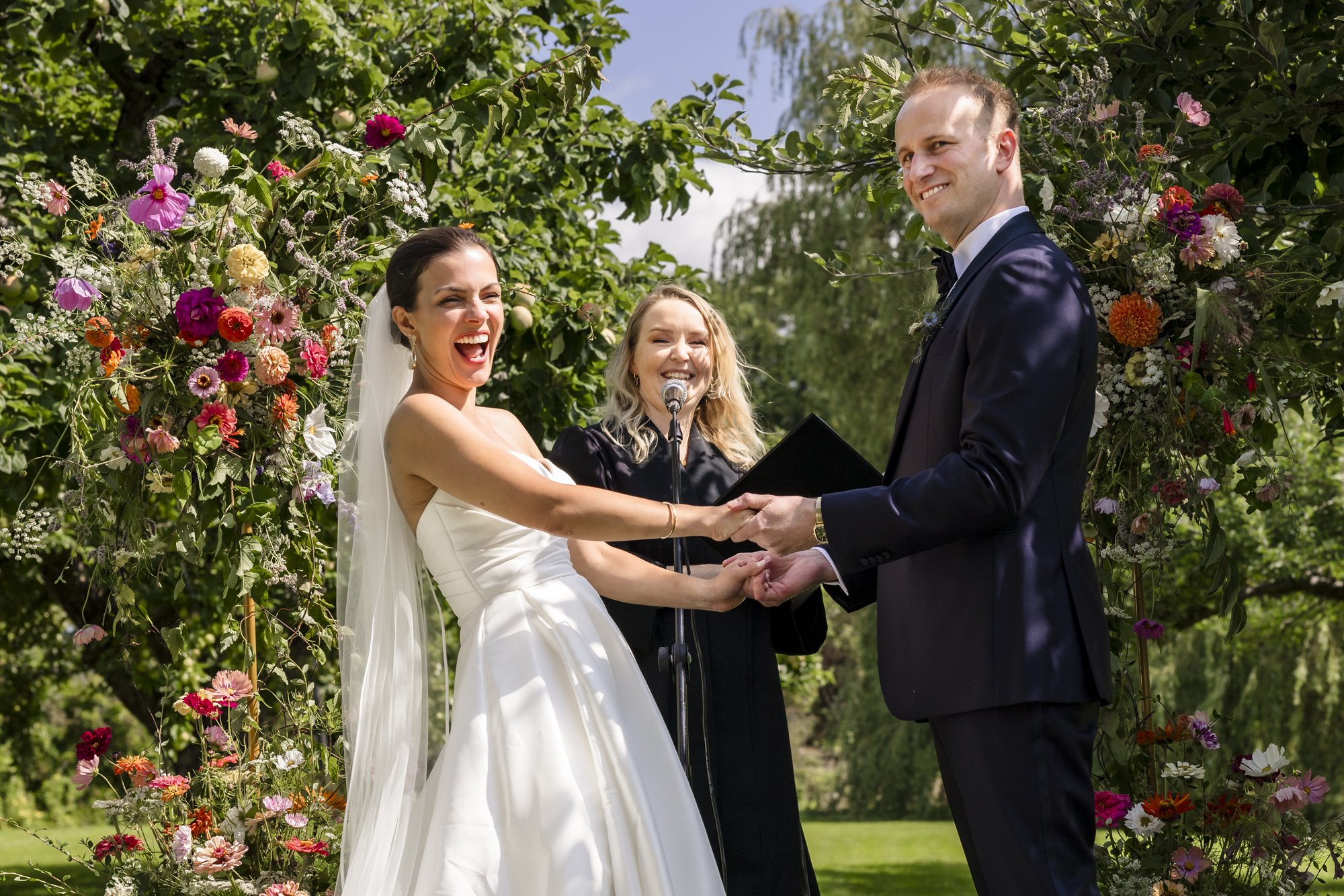 Et par gifter seg utendørs under en blomsterdekorasjon med to personer i formell antrekk, og en kvinnelig offisiant.