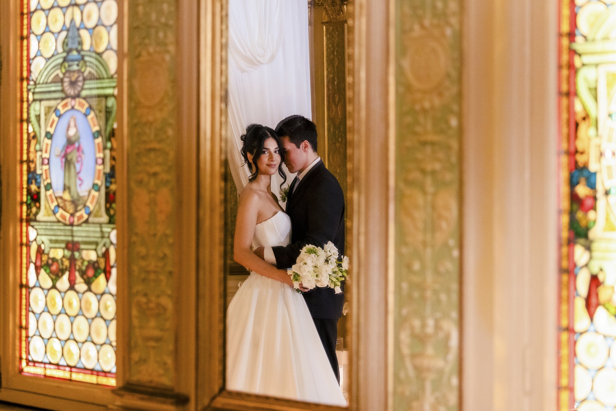 Et brudepar står nær hverandre i en hall med fargede glassmalerier og gule malerier på veggene, de ser på hverandre og holder en blomsterbukett.