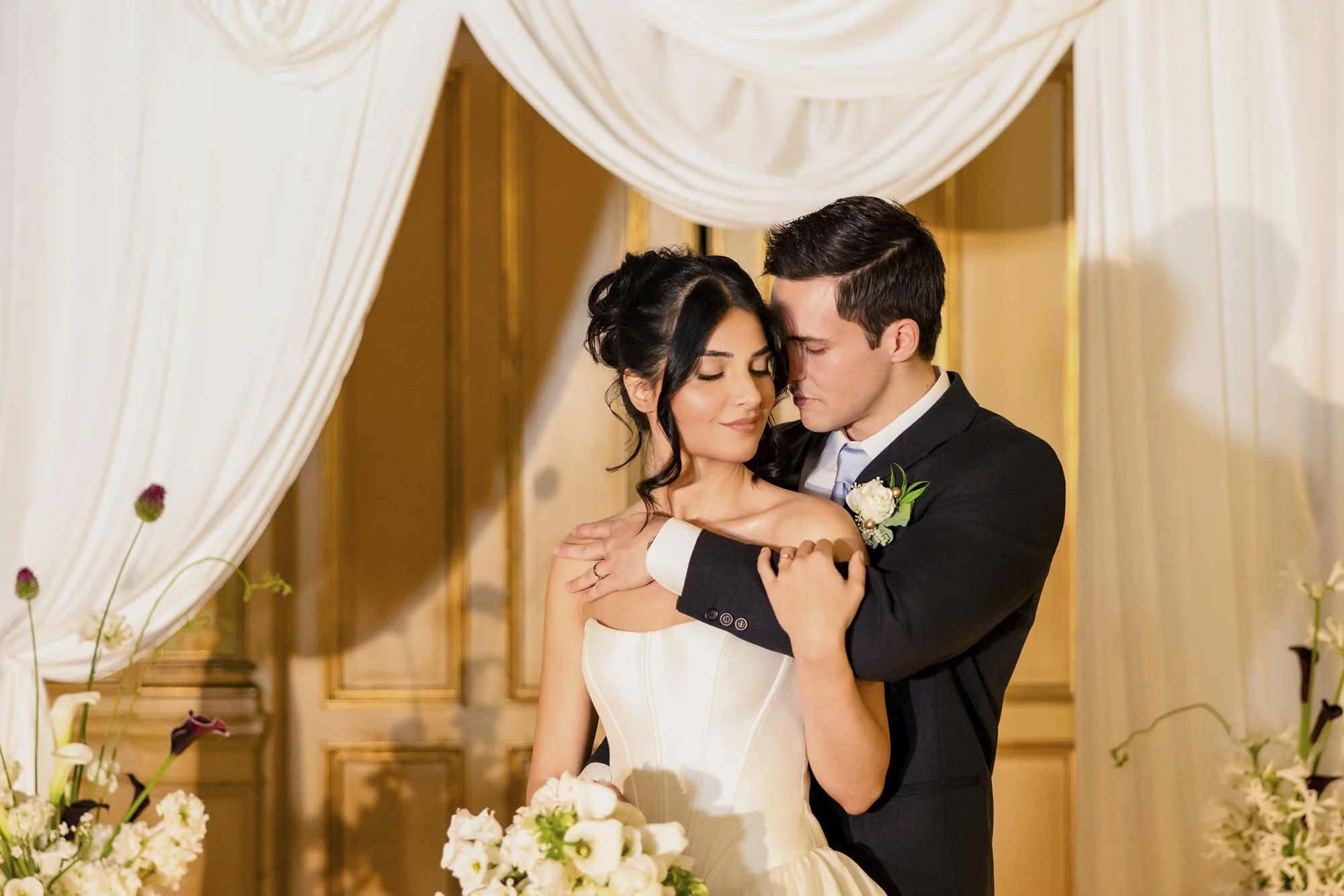 Et brudepar i bryllupsklær omfavner i et romantisk øyeblikk under seremonien, omgitt av hvite blomster og gardiner.