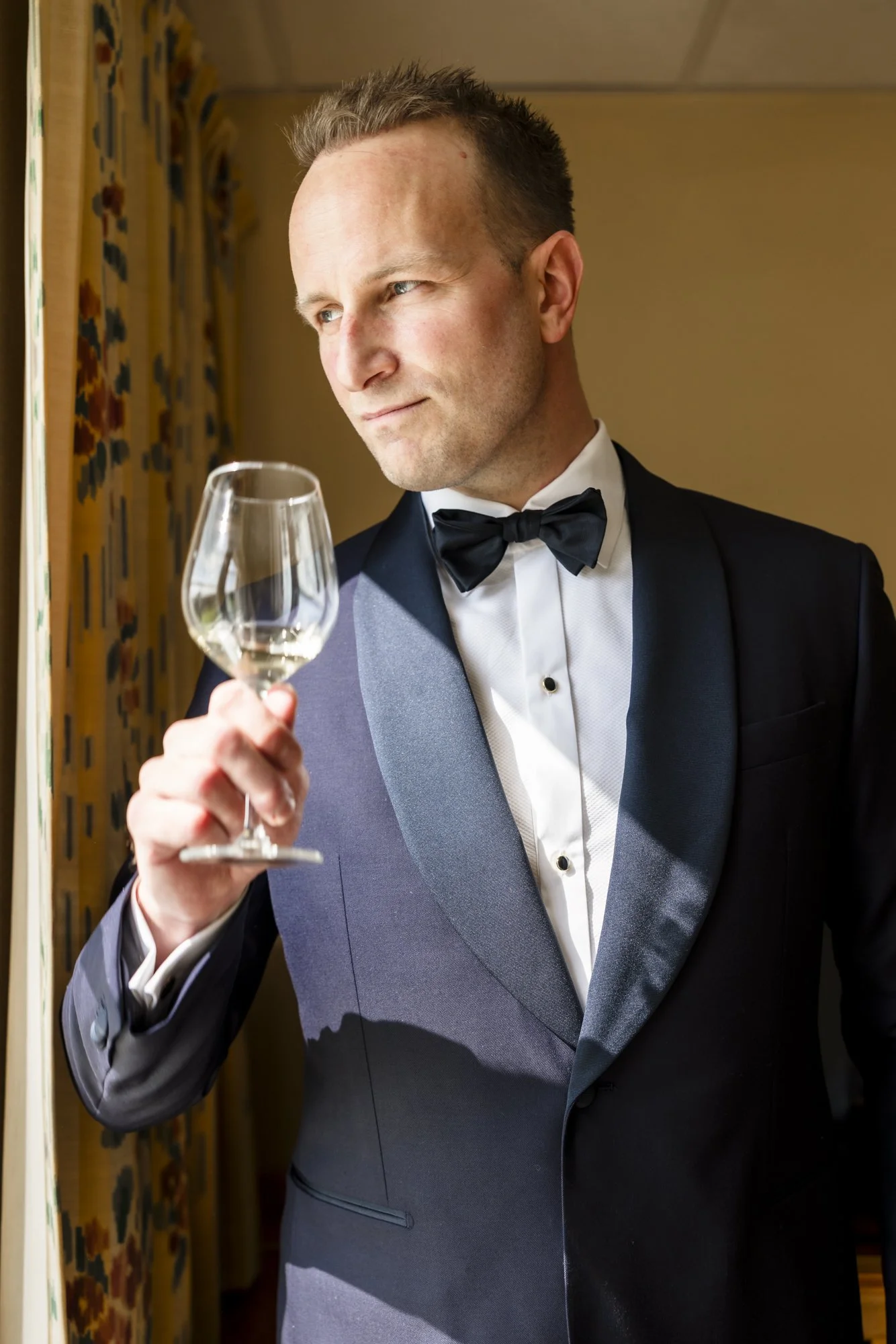 En mann i svart smoking med hvit skjorte og svart sløyfe ser ut av vinduet mens han holder et vinglass med hvit vin.