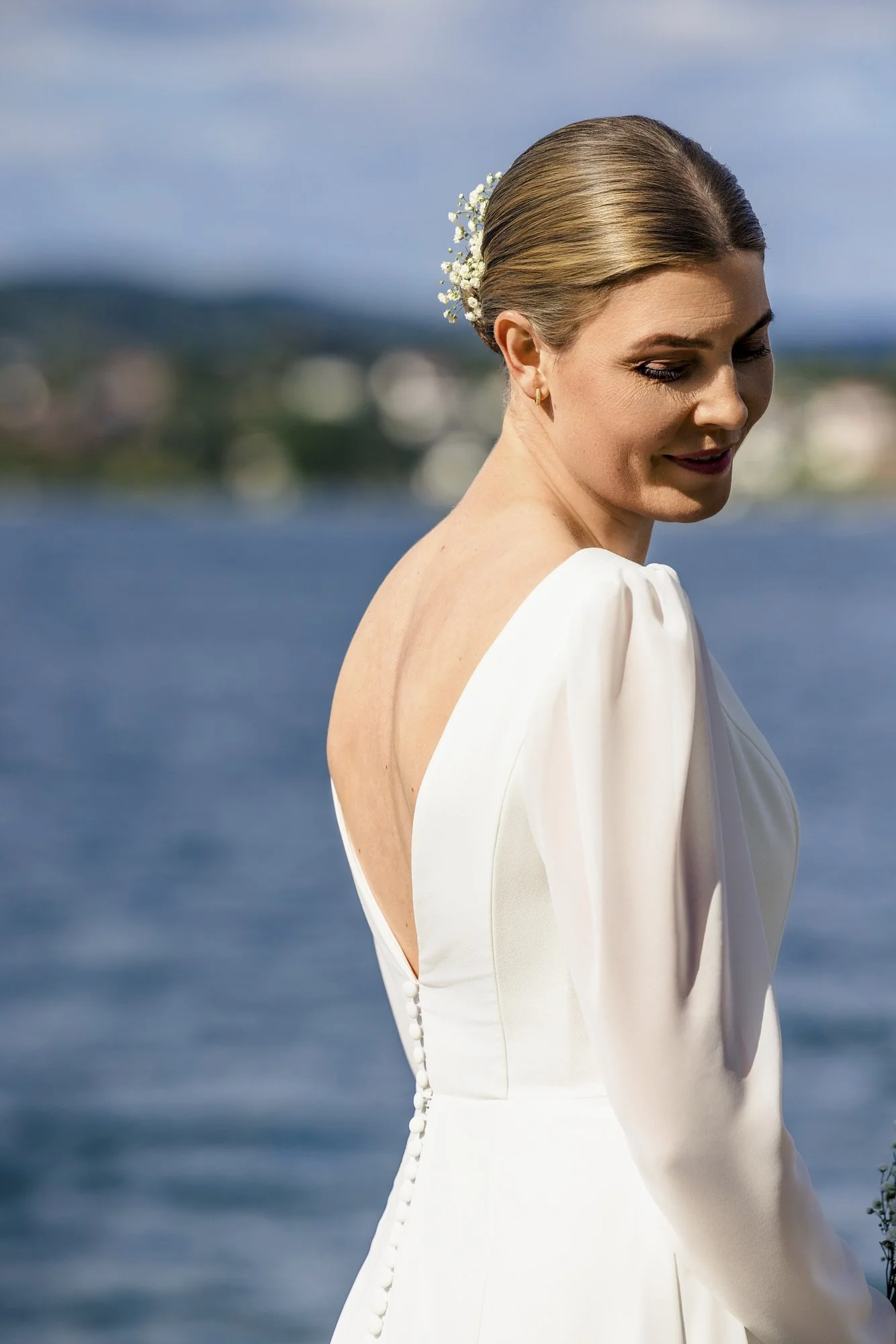 Kvinne i hvit brudekjole ved vannet, har hår pyntet med blomster, smiler forsiktig, med bylandskap i bakgrunnen.