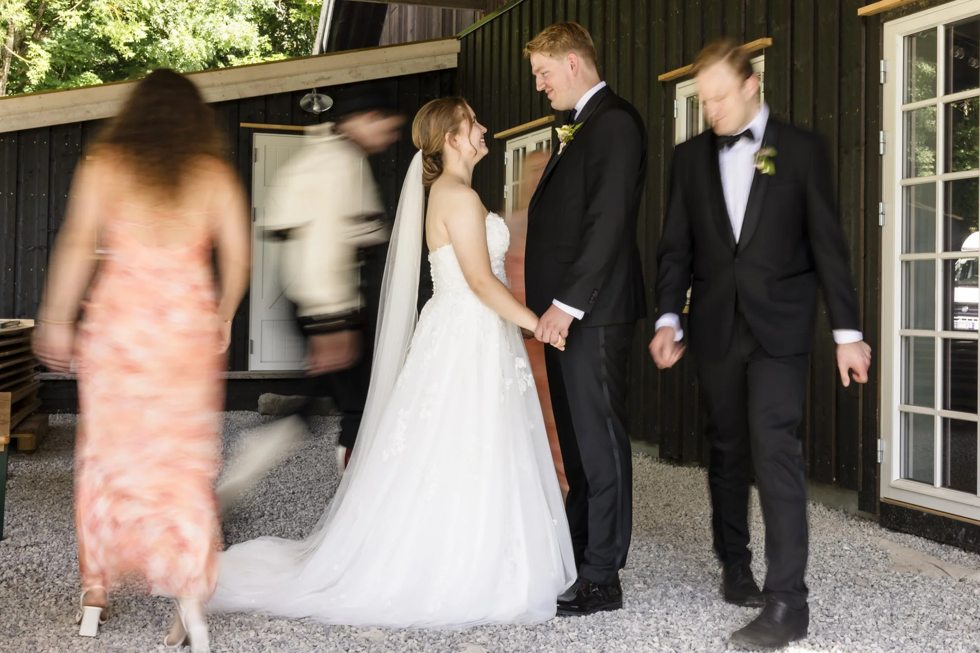 Brudepar som holder hender og ser på hverandre, omgitt av brudesvenner under en bryllupsfeiring utendørs, med hekk og bygning i bakgrunnen.