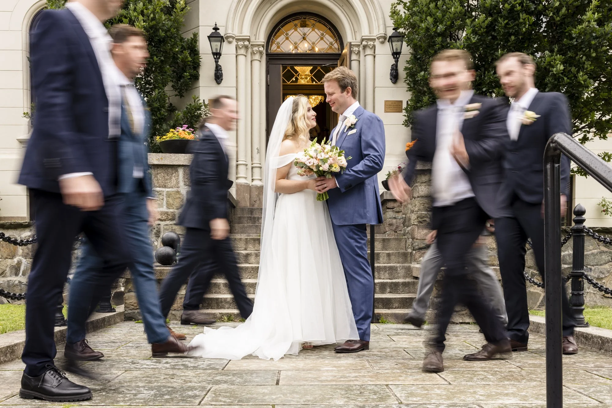 En brud og en brudgom står sammen foran inngangen til en bygning, omgitt av venner eller familie i fint antrekk, noen av dem er i bevegelse. Bruden holder en bukett blomster, og paret ser på hverandre med smil. Bakgrunnen viser en lys bygning med trapp og utemiljø.