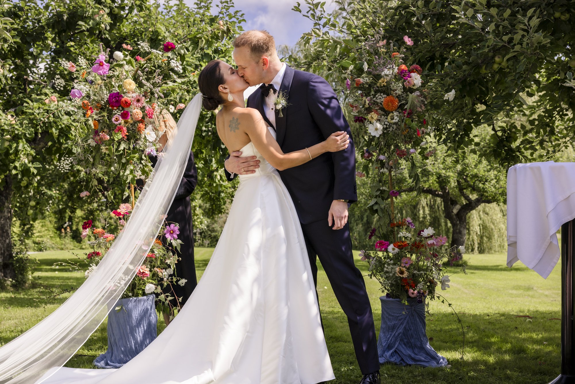 Et brudepar kysser under en blomsterdekorasjon utendørs under bryllup.