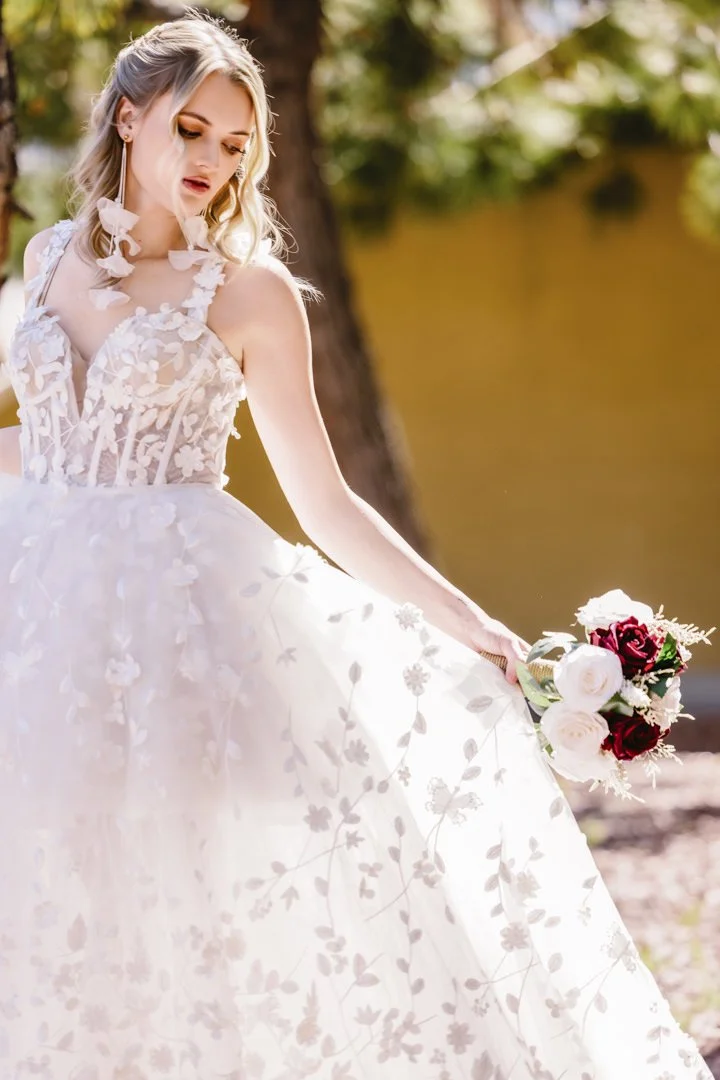 En ung kvinne i en brudekjole holder en blomsterbukett utendørs ved daggry, omgitt av sollys og trær.