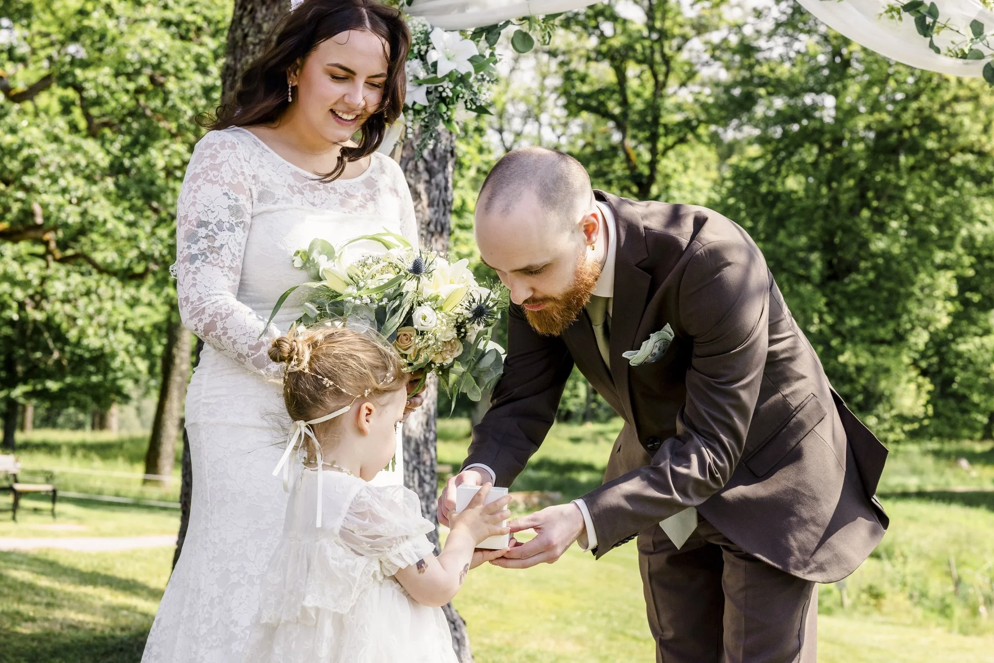 En brud med brunt hår og en brudgom med skjegg deler en gave med en liten jente i en utendørs vielse.