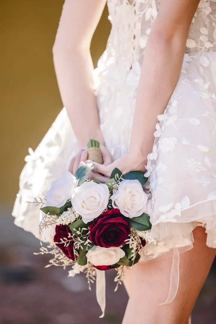 En person i en hvit brudekjole holder en bryllupsbukett med røde og hvite roser.