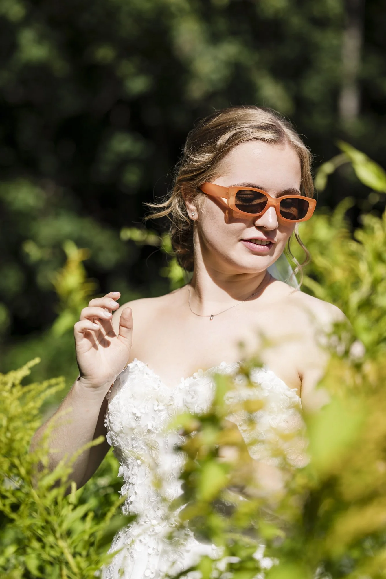 Kvinne med brunt hår, iført hvit kjole med blomsterdetaljer, poserer utendørs blant grønt buskverk, iført oransje solbriller og smilende.
