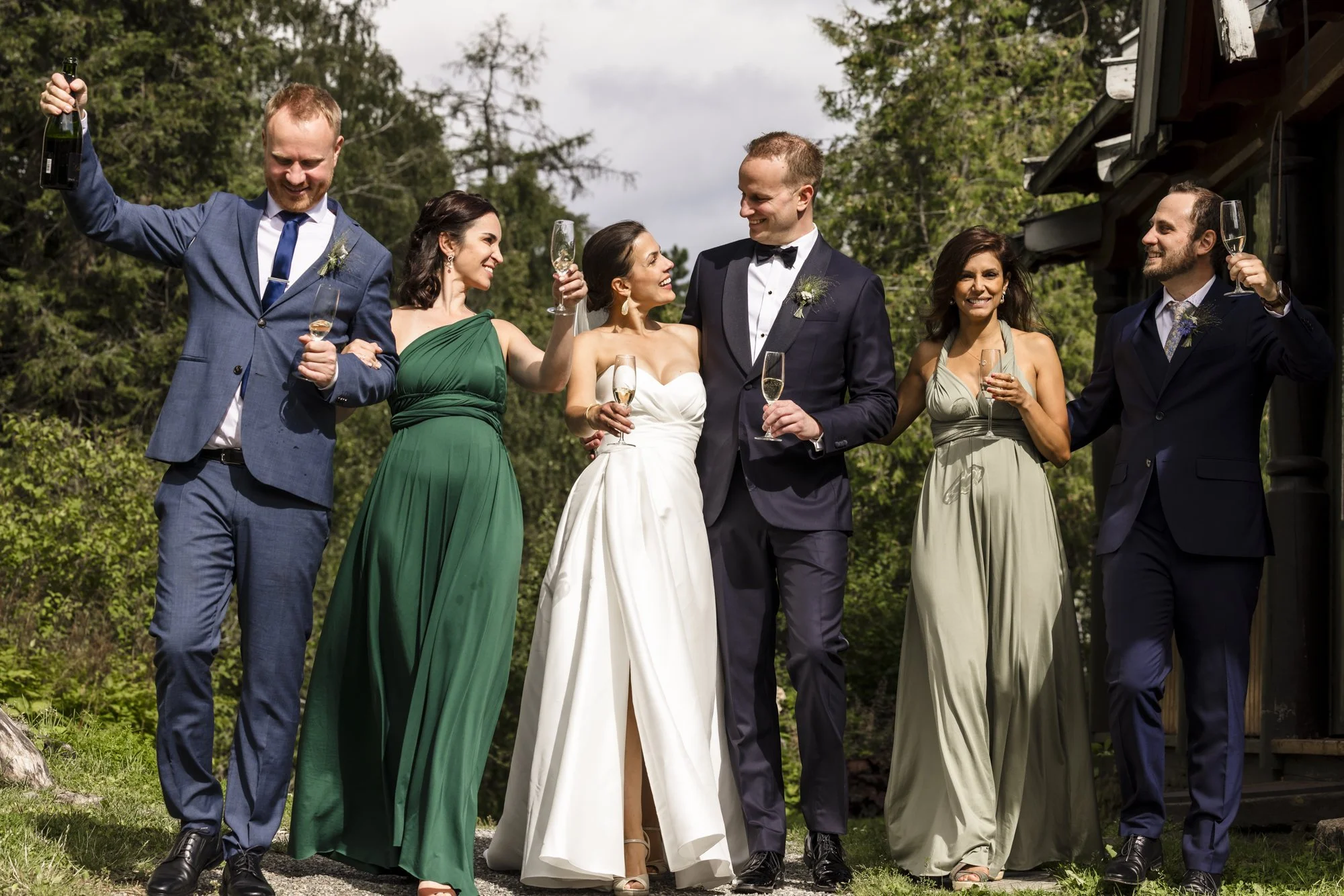 Syv personer som feirer med champagne, to er i brudekjole og dress, ute i naturområde.