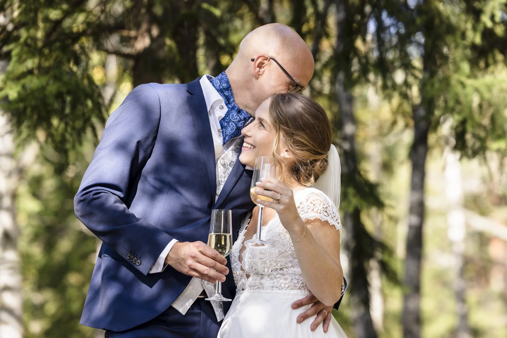 Et bryllupsbilde av et par som smiler og holder champagneglass, utendørs blant trær.