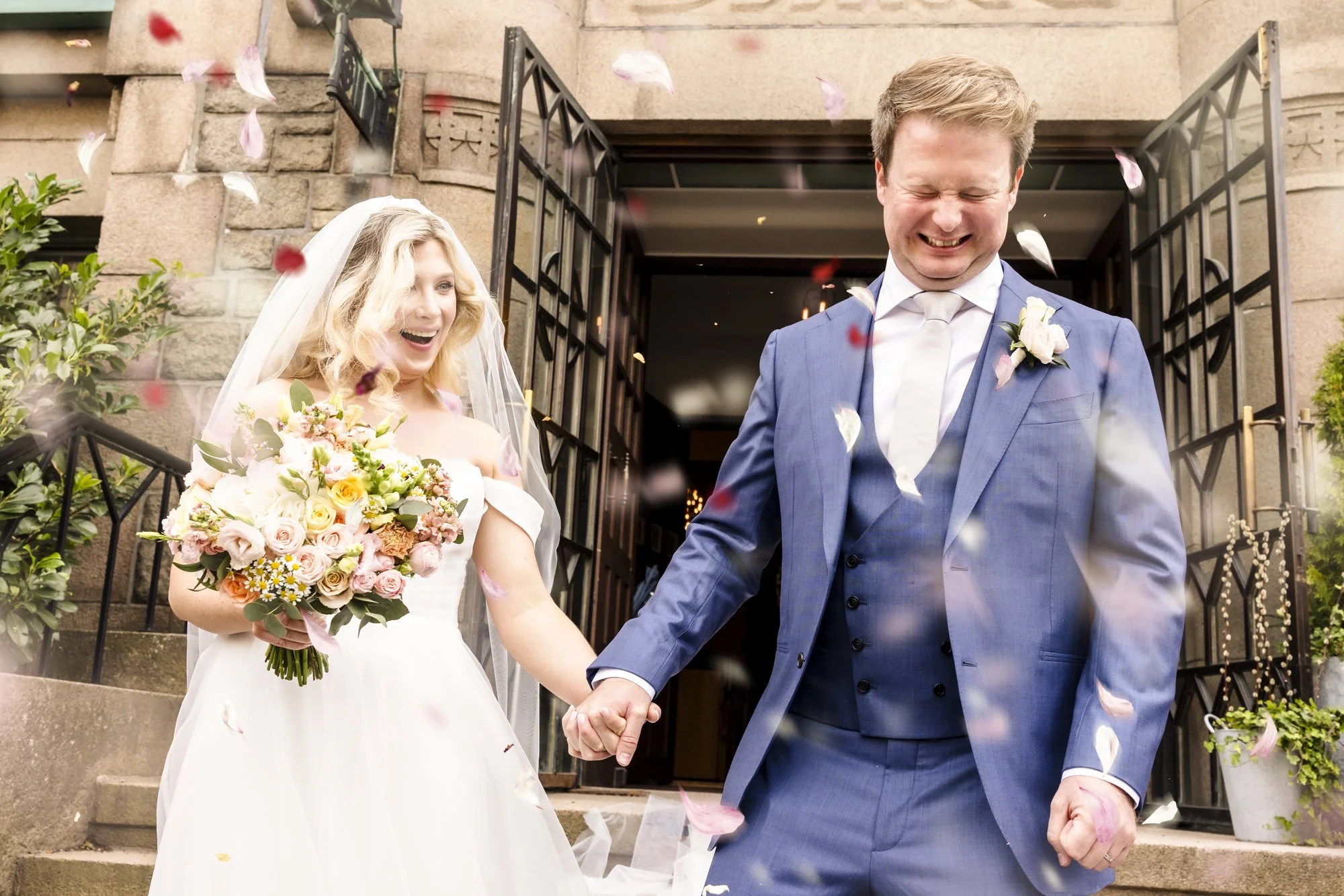 Et ektepar på bryllupsdagen, smilende og holder hender, står foran en bygning med åpen dør, med blomster og blomsterblad som faller ned rundt dem.