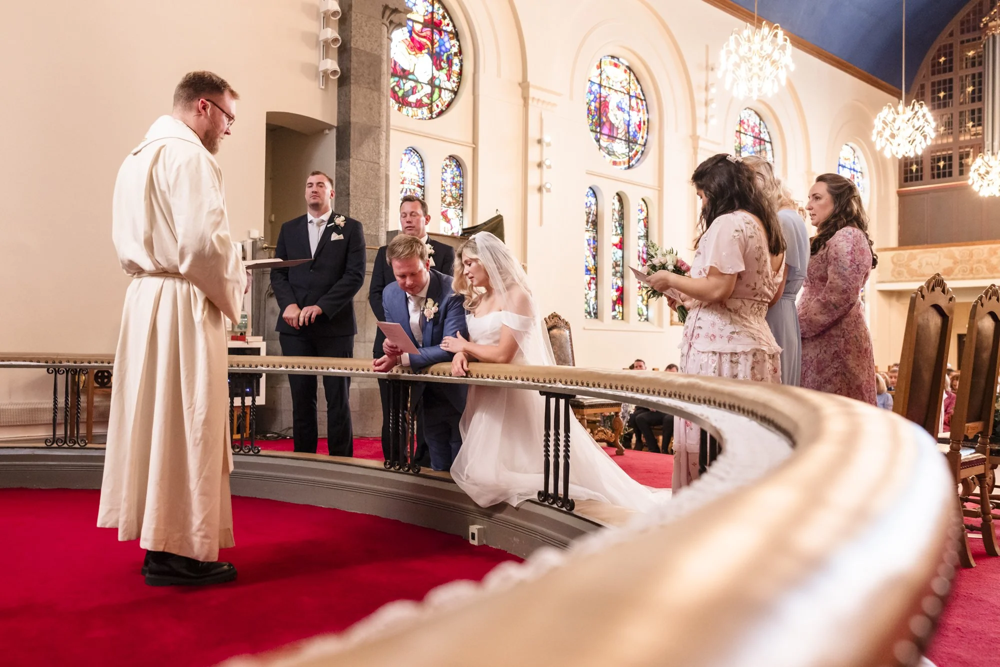 Bryllupsseremoni i en kirke med presten som står foran paret som kler seg i vielsesklær, mens bruden og brudgommen og vitner står bakerst og ser på. Kirken har store fargerike glassmalerier og pene lysekroner.