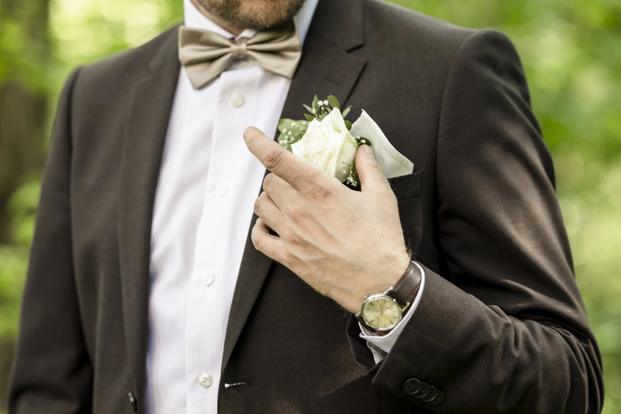 Man i brun dressjakke med lysfarget skjorte og beige sløyfe, holder en hvit blomsterlapp i brystlomma.