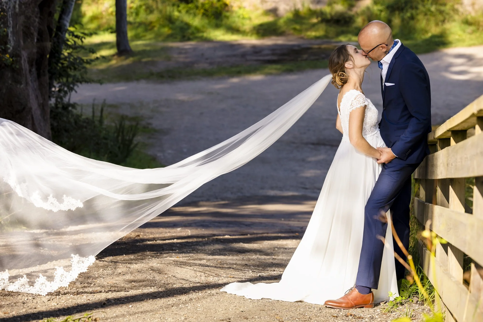 Et brudepar kysser under en bue av tårefast brudekappe, med dekket av trær og en sti i bakgrunnen.