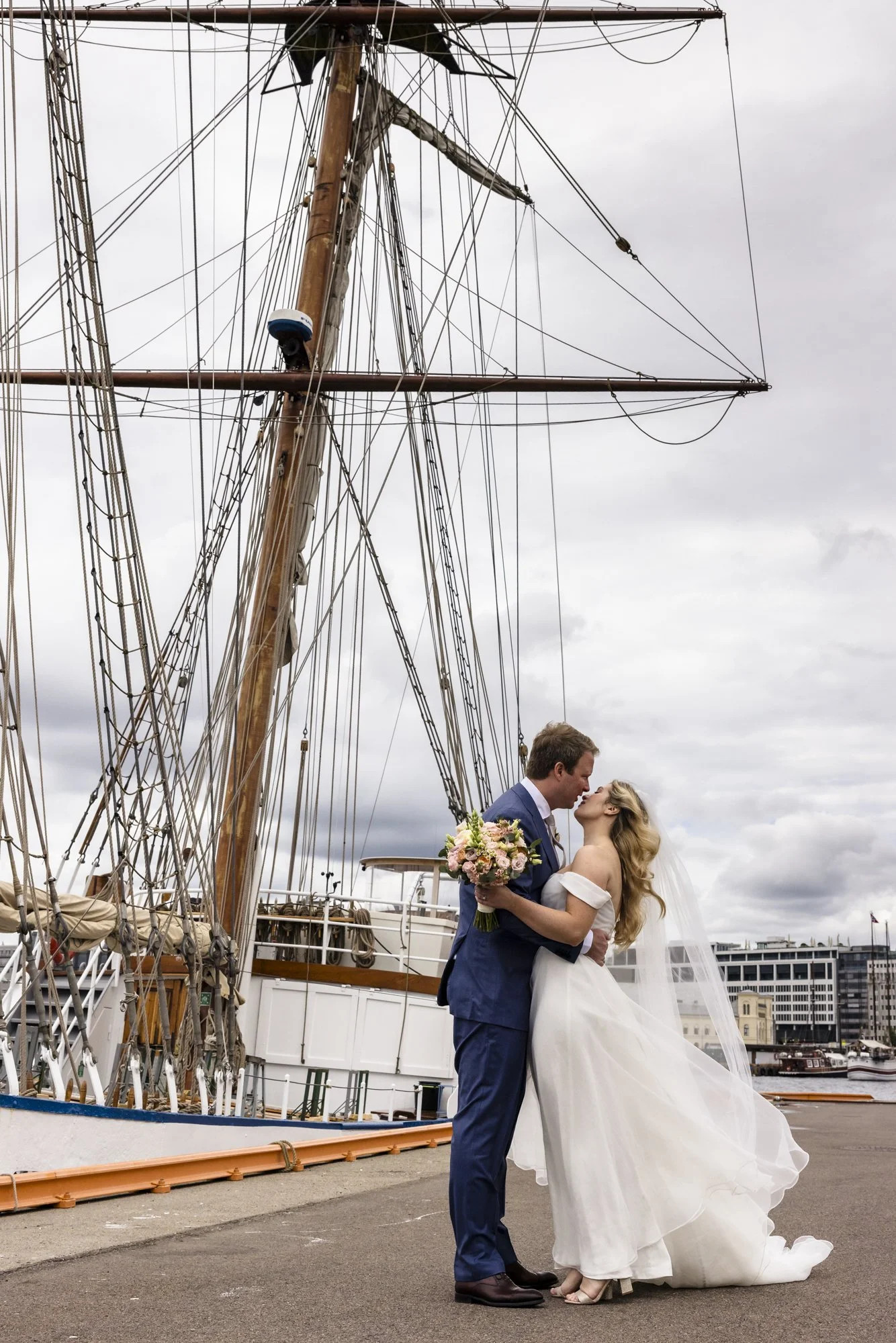 Et brudepar står foran en stor seilskute, de er i en romantisk kyss, bruden holder en bukett blomster, og de er utendørs ved havnen under en overskyet himmel.