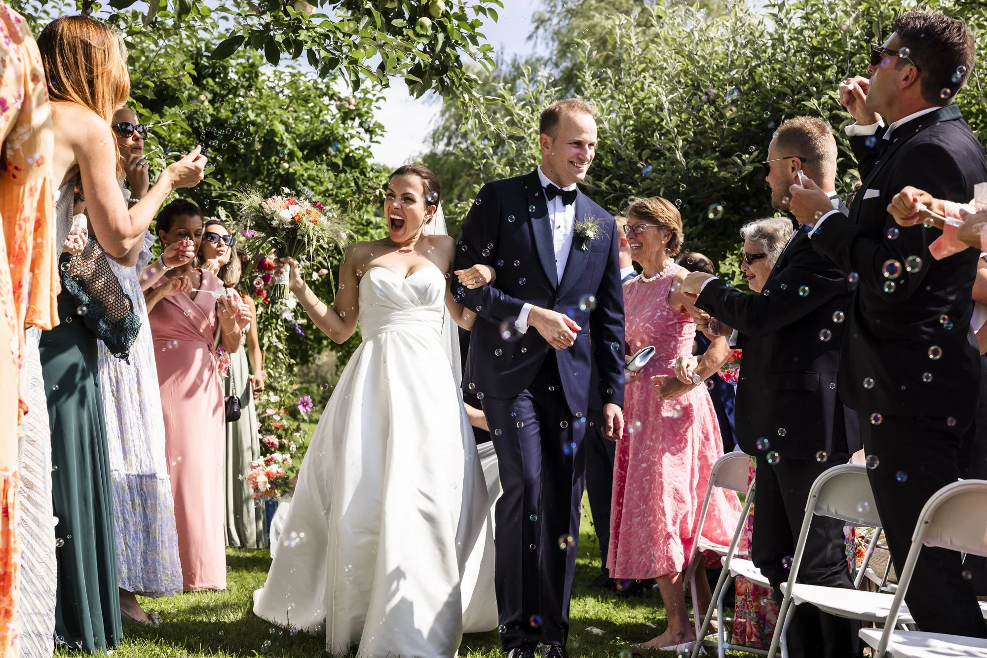 Et brudepar går ned rampen etter vielsen, omgitt av venner og familie som feirer, omgitt av blomster og grønt. Både bruden og brudgommen smiler, og det er såpebobler i luften.