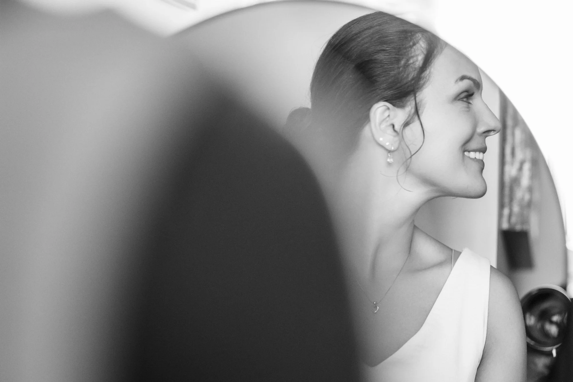 En smilende kvinne med oppsatt hår, iført en hvit kjole og smykker, sett fra siden. Bilde i svart-hvitt.