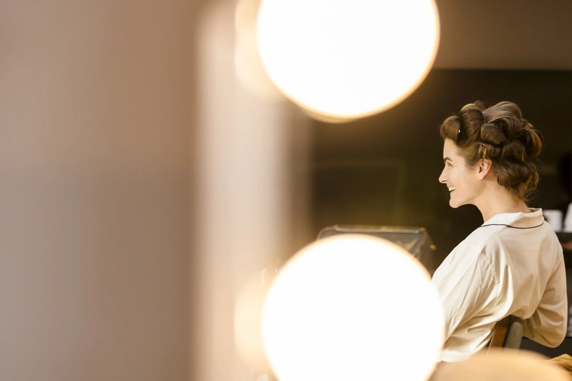 Kvinne med voluminøse, rullede hår, smilende og sett fra siden, sitte i et rom med varme lys.