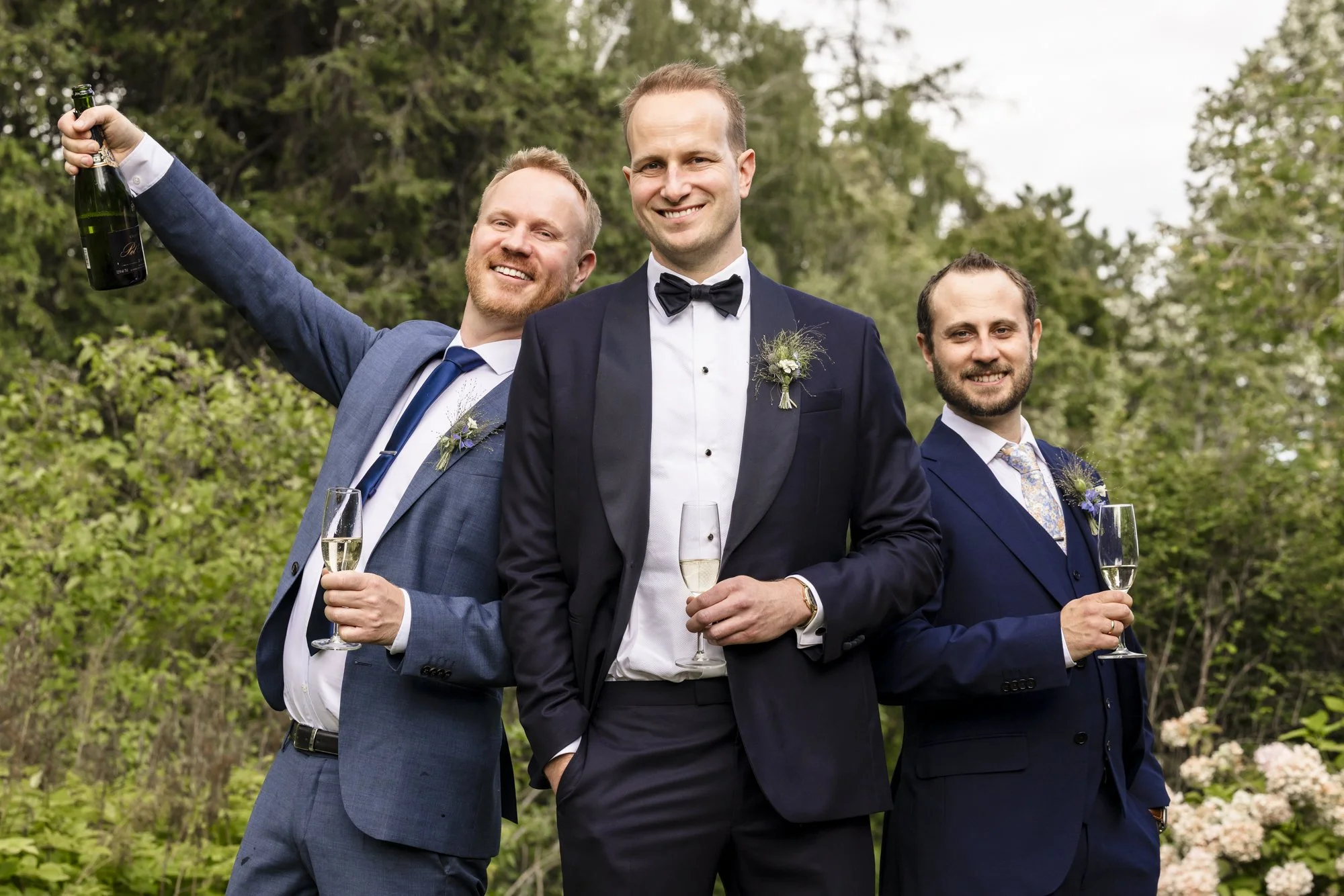 Tre menn i formelle dresser, to holder champagneglass, den midterste har på seg smoking og bowtie, alle smiler, utendørs med grønne trær i bakgrunnen.