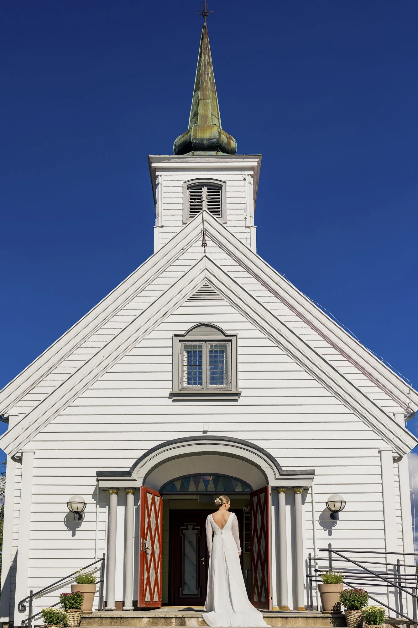 En kvinne i en hvit brudekjole står foran den åpne inngangen til en hvit kirke med høyt spiret tårn under en klar blå himmel.