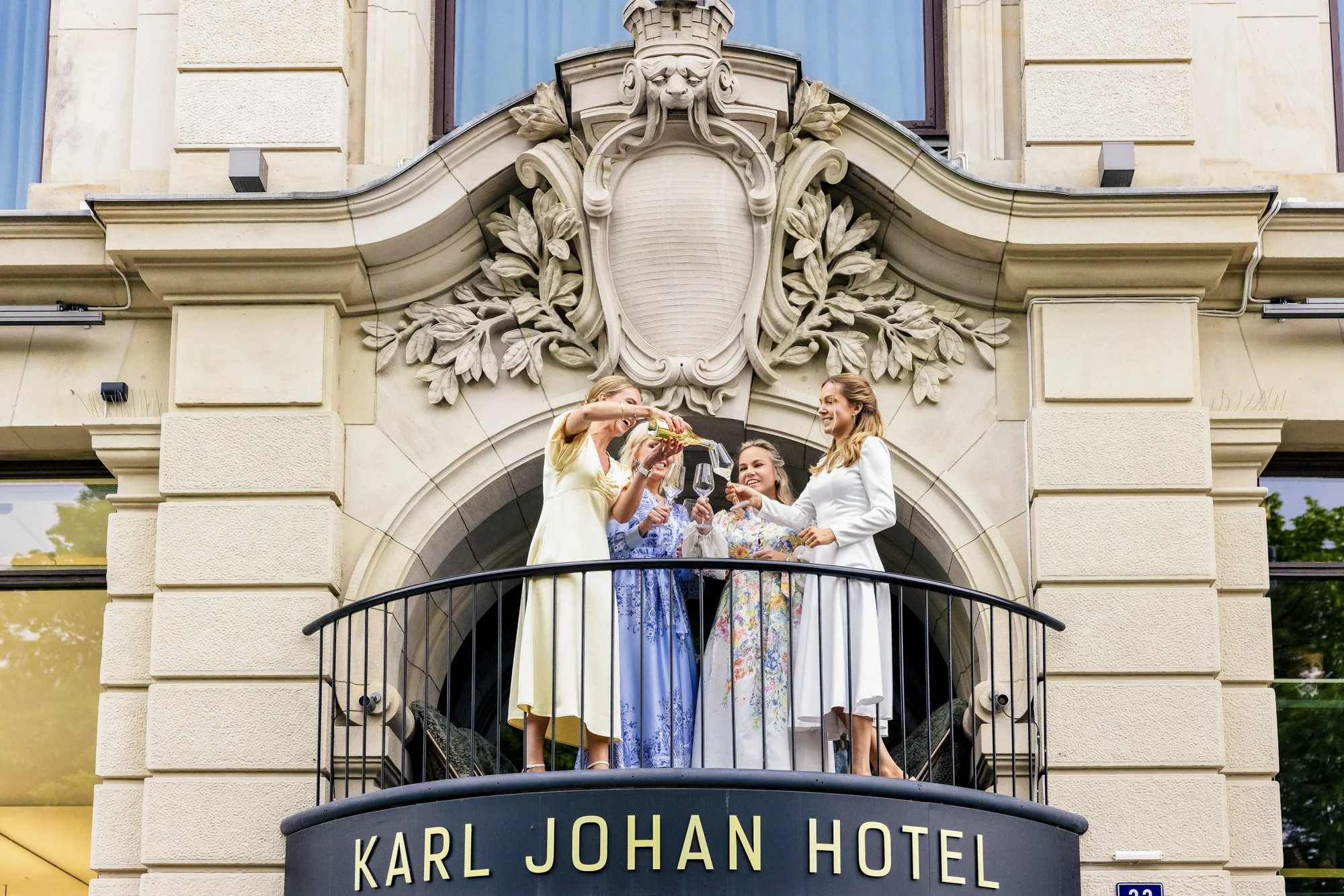 Fire kvinner i elegante kjoler på balkongen foran Karl Johan Hotel, skåler med champagne, smiler og har en festlig anledning.