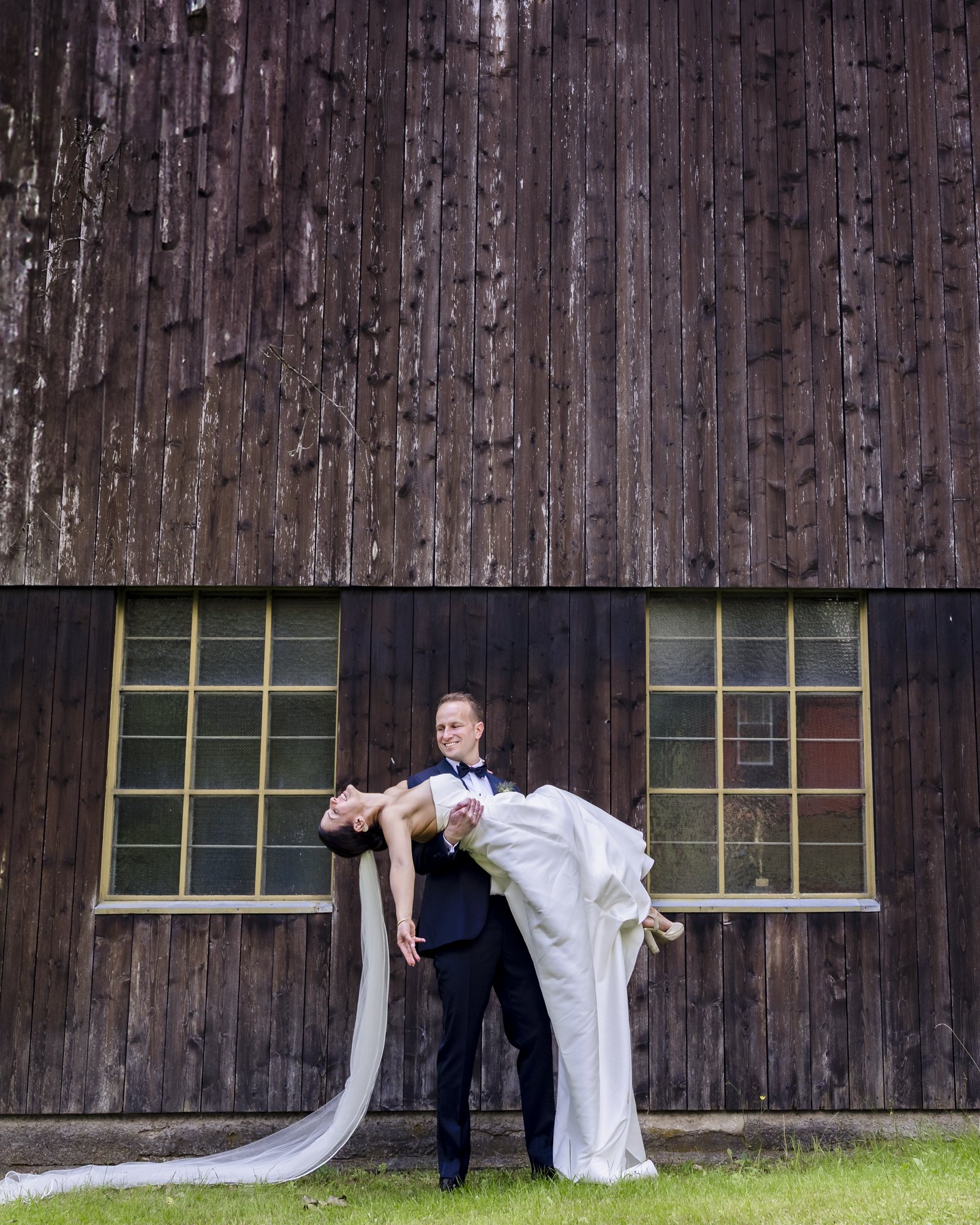 En brud og en brudgom står foran en gammel, mørk trebygning med to store vinduer, og brudgommen bærer bruden i armene mens de smiler.